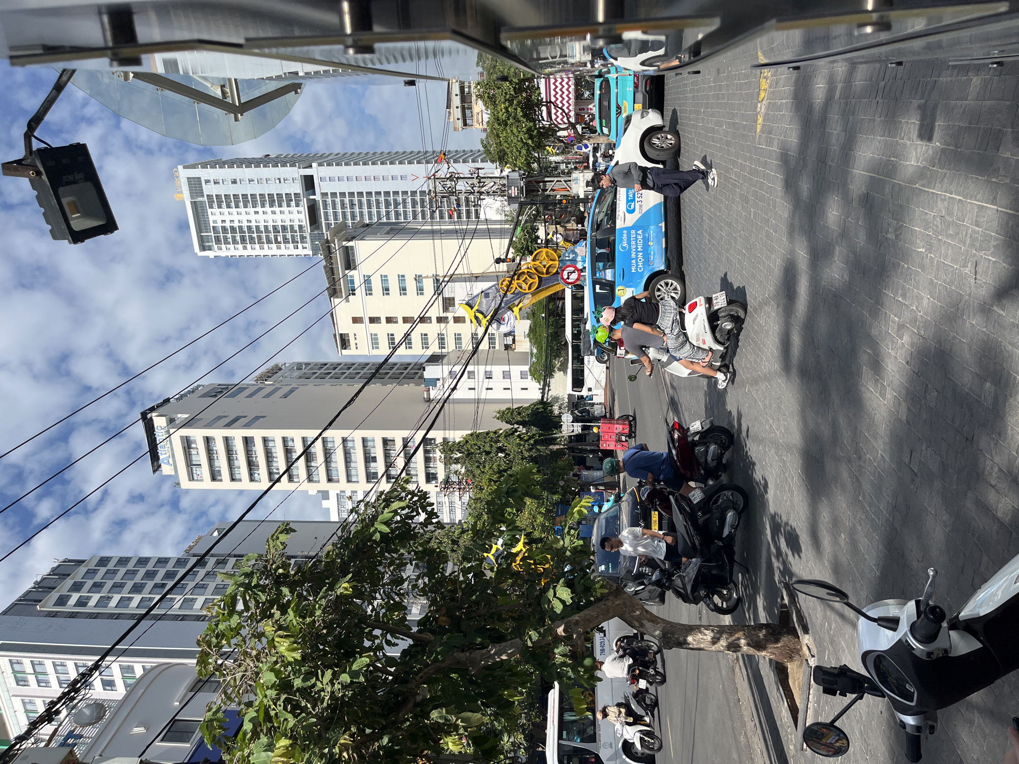 This image captures a street scene in Nha Trang, Vietnam, on a partly cloudy day. The foreground features the front of a scooter, implying the vantage point of someone on a motorbike. To the left, a white bus is parked, with a person on a scooter visible beside it. Further down the street, a line of cars and motorbikes are navigating the road. Prominent in the midground is a blue and white minivan with "Midea" branding, suggesting it's a company vehicle. A group of people on motorbikes are in motion or stopped. Tall buildings dominate the background, characteristic of an urban environment. The road is paved with interlocking grey stones, and a sidewalk with a tree is present on the left. Overhead, a complex network of electrical wires crisscrosses the sky.
