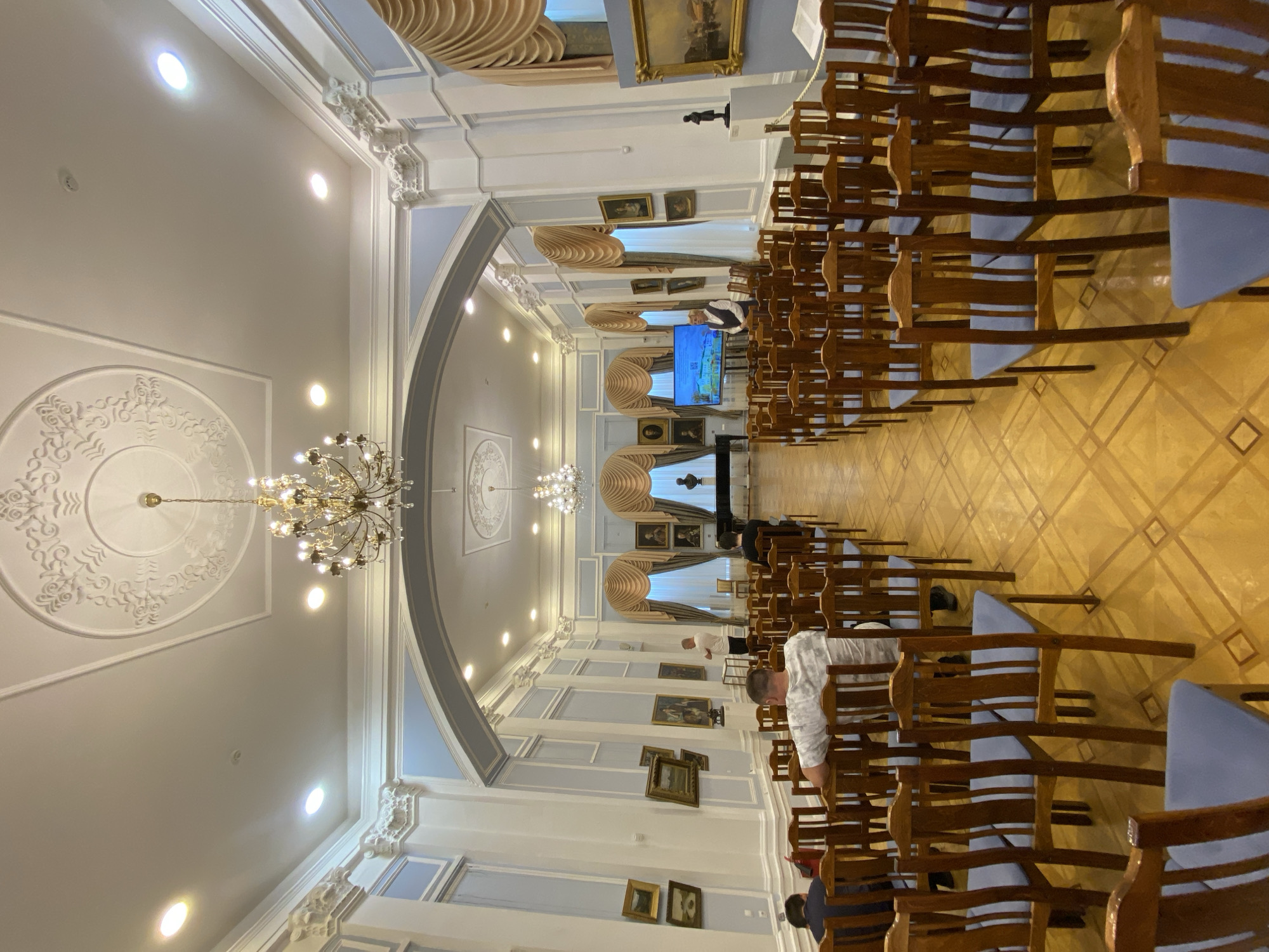 The image shows an indoor event space with rows of empty wooden chairs with blue seats arranged on a polished wooden floor. The room is elegantly decorated with high ceilings, ornate moldings, and a large chandelier. Paintings are displayed on the walls, and there are large windows with draped curtains. At the front of the room, there is a stage area with a screen displaying an image of a building, a grand piano, and a bust. Several people are visible, including a speaker standing near the screen and a few attendees seated in the chairs. The overall impression is that of a cultural or educational venue, possibly a museum, lecture hall, or art gallery. The location is identified as the Museum of Fine Arts in Petrozavodsk, Russia, a city known for its cultural institutions.