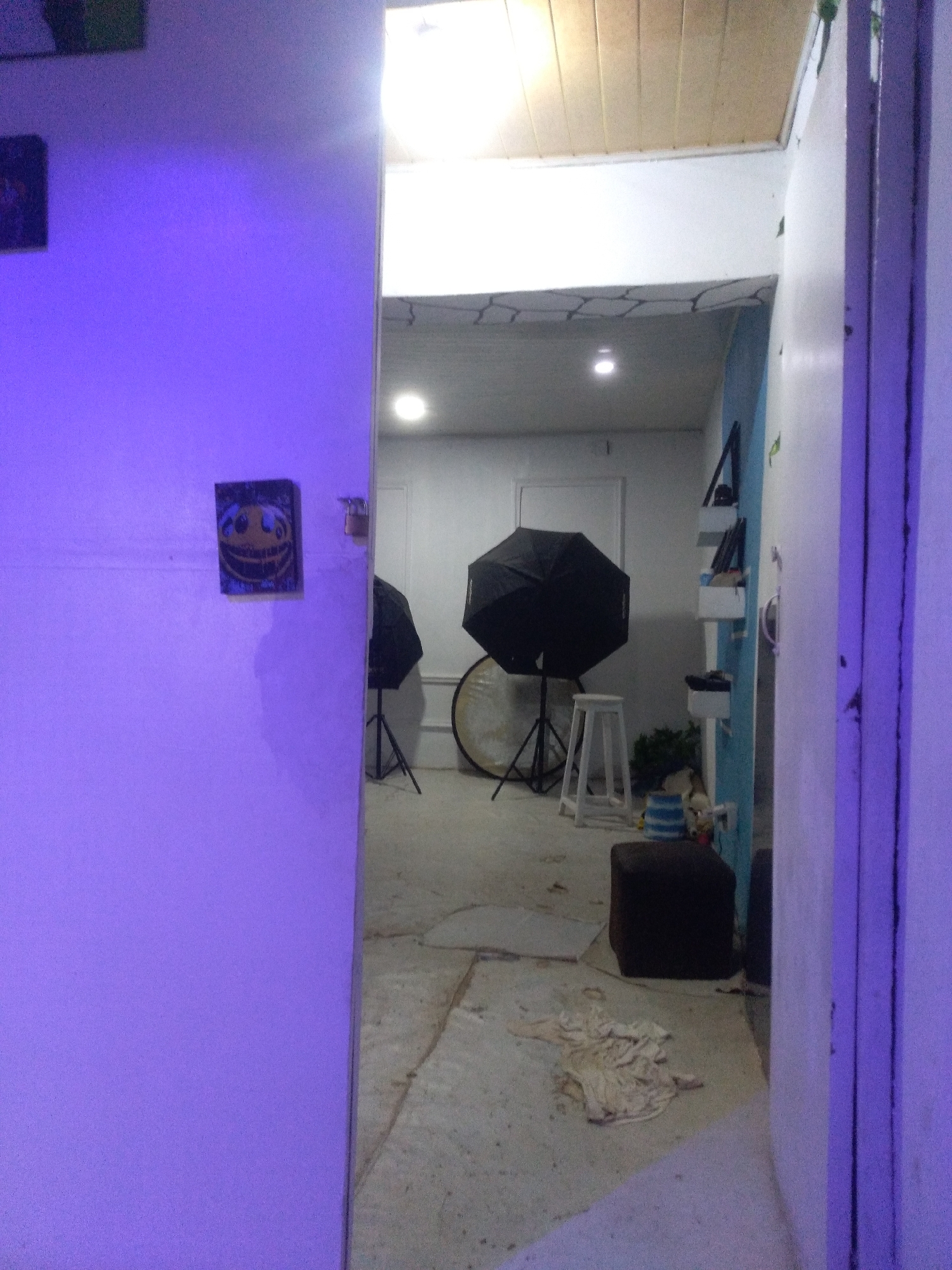 The image captures an interior view, looking through a doorway from a room bathed in purple light into an adjacent, brighter space that appears to be a photography studio or creative workspace.
In the immediate foreground on the left, a purple-lit wall dominates, with a small rectangular picture or painting featuring a stylized, possibly grinning or unsettling, smiley face hanging on it. Above this, another dark framed picture is partially visible in the upper left corner. The doorway on the right shows a white door frame, also illuminated with purple light.
Beyond the doorway, the second room is brightly lit with overhead lighting, some of which is visible, and features a light-colored ceiling, part of which has wooden paneling. The walls in this room are primarily white, with one section on the right painted a light blue and fitted with several open white shelves or storage bins.
Within this studio space, there are several pieces of photography equipment: two black octagonal softboxes or umbrellas are set up on stands, and a large circular silver reflector rests on the floor nearby. A white four-legged stool stands between the softboxes. On the floor in the foreground of the studio, a dark brown ottoman or small stool is visible, along with a crumpled, light-colored cloth. Some greenery or plants are partially visible behind the white stool and near the blue wall. The floor in this area appears to be concrete or a similar light-colored, somewhat rough surface. No people or legible text are visible, and no specific activity is taking place. The overall impression is that of a prepared, but currently unoccupied, creative space.