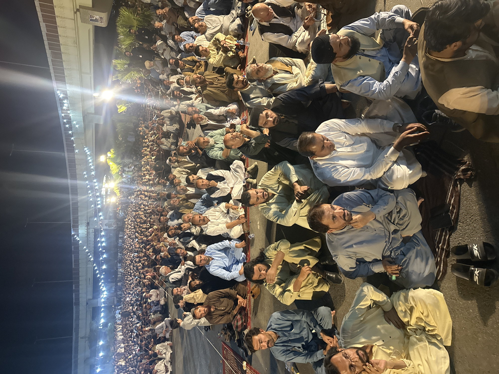 An outdoor crowd of men is gathered at night in Quetta City Tehsil, Pakistan, under an elevated highway structure adorned with string lights. The majority of the individuals are seated on the ground, some on rugs and mats, while others are directly on the pavement. They are predominantly dressed in traditional South Asian attire, consisting of shalwar kameez and vests. The group appears to be attentive, with many looking in the same direction, suggesting a communal gathering or event. The lighting is a mix of overhead streetlights, the string lights, and some spotlights creating bright areas and shadows. The atmosphere seems solemn or focused.