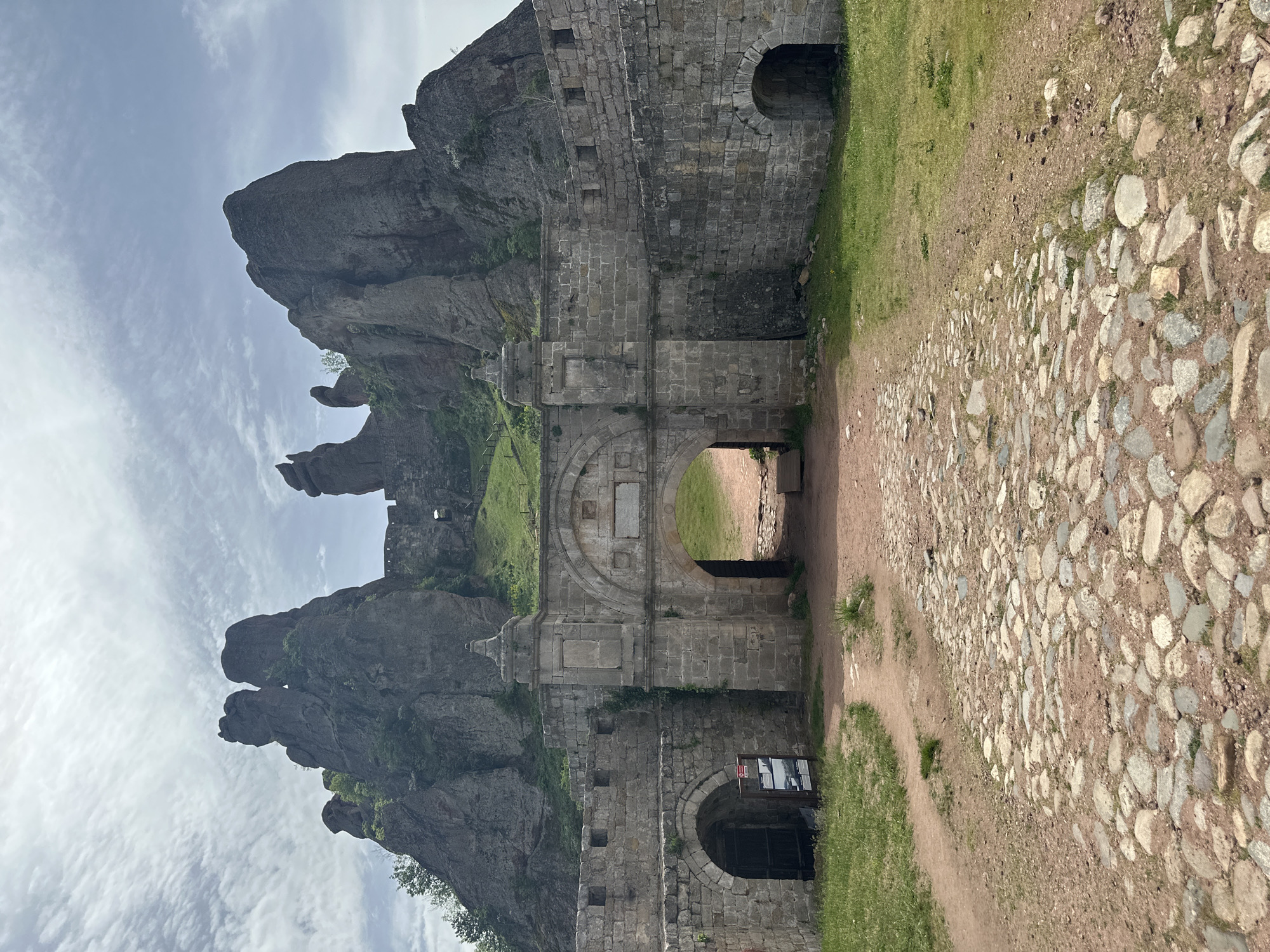 The image depicts a gate in Belogradchik Fortress, situated in Belogradchik, Bulgaria. The fortress is constructed of stone and integrated with the surrounding rock formations. The gate features arched entrances and is flanked by stone walls. The foreground shows a pathway made of cobblestones and dirt. Green grass grows along the edges of the path. In the background, there are large rock formations that dominate the landscape. The sky is partially cloudy.