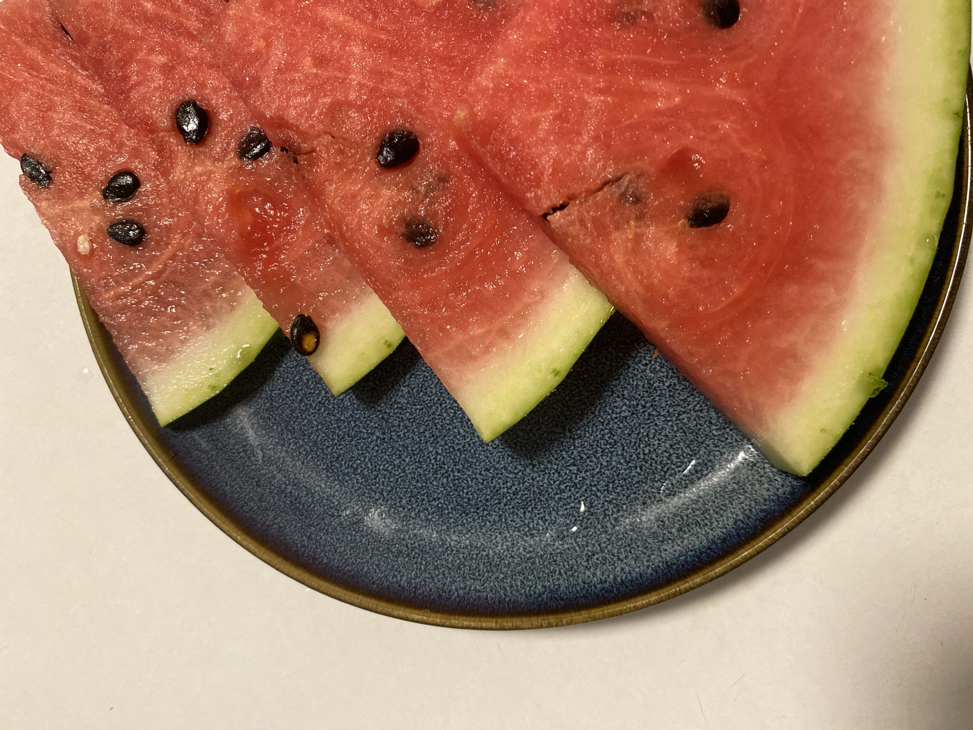 The image shows a partially visible blue speckled plate with a brown rim, on a white surface. On the plate are four slices of watermelon with black seeds and rind. The slices are arranged in a slightly overlapping manner. The location is not discernible from the image.