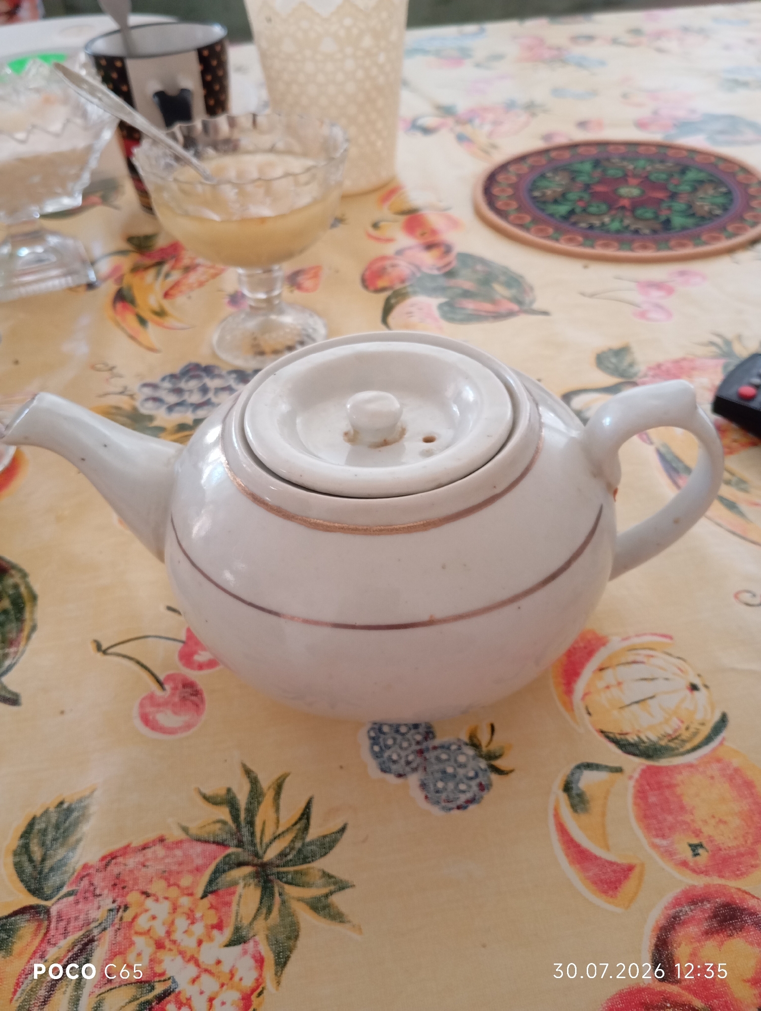 The image shows a white teapot with a gold trim on a tablecloth with a fruit pattern. The teapot is in the center of the frame. In the background, there are two glass dishes, one containing a light-colored liquid and the other containing a white granular substance, and a mug. There is also a decorative coaster on the tablecloth to the right of the teapot. The scene appears to be set indoors, likely on a table. No people are visible in the image. The date and time stamp "30.07.2026 12:35" is present in the bottom right corner, along with "POCO C65" in the bottom left. The location of Beruniy, Uzbekistan, is not indicated in the image itself.