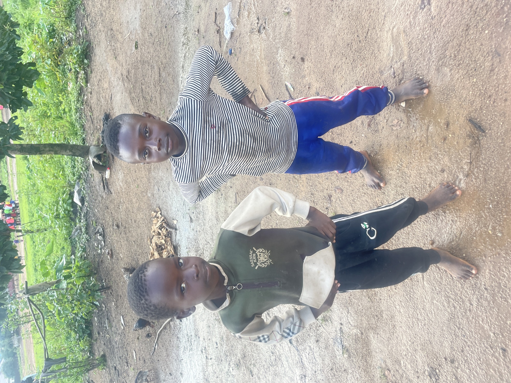 Two young boys are standing on a dirt ground in what appears to be an outdoor area with some vegetation in the background. The boy on the left is wearing a green and beige long-sleeved shirt with a white pocket and black pants. He has his hands on his hips and is looking at the camera. The boy on the right is wearing a striped long-sleeved shirt and blue pants with red stripes. He also has his hands on his hips and is looking at the camera. The ground is wet and muddy, with some scattered debris. In the background, there is more vegetation, a tree, and a glimpse of a group of people in colorful clothing in the distance. This scene is consistent with a rural or semi-rural setting in the city of Jos, Nigeria, possibly after rainfall.