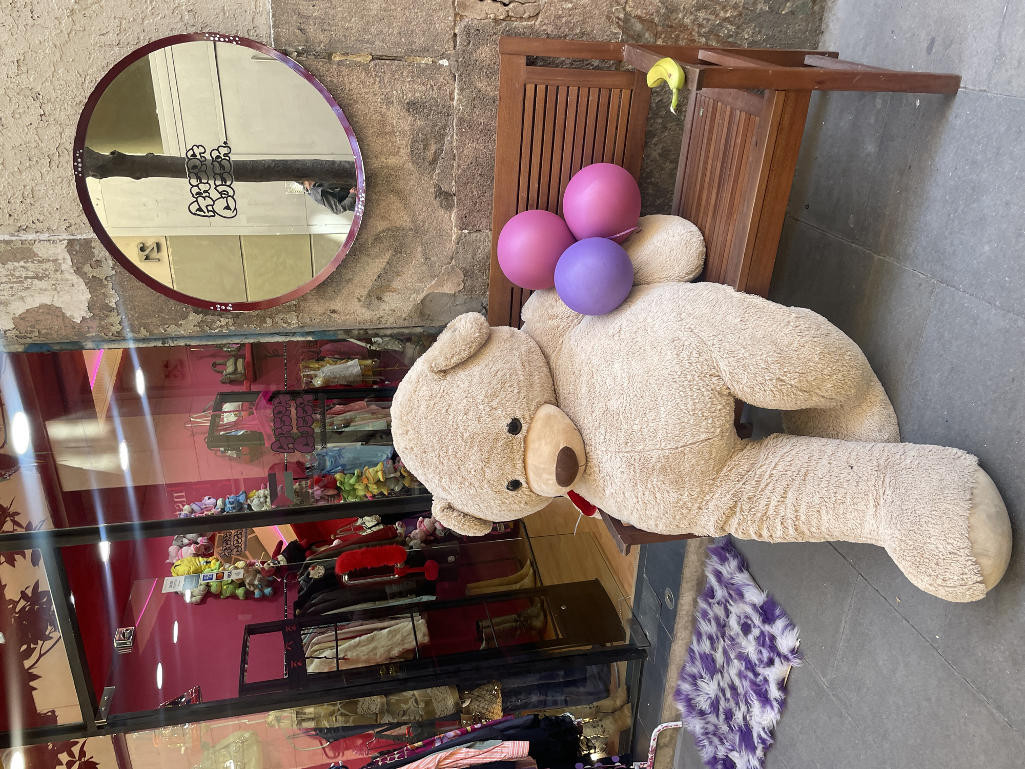 The image shows a large, beige teddy bear lying on a small wooden bench outside a shop in Barcelona, Spain. Three balloons, two pink and one purple, are nestled against the bear's back. A round mirror with a maroon frame hangs on the stone wall behind the bench. The shop's display window is visible, showcasing various items of clothing and accessories. A small, purple and white rug sits on the pavement near the bear's leg. A banana peel is draped over the back of the bench. The scene is bathed in natural light.