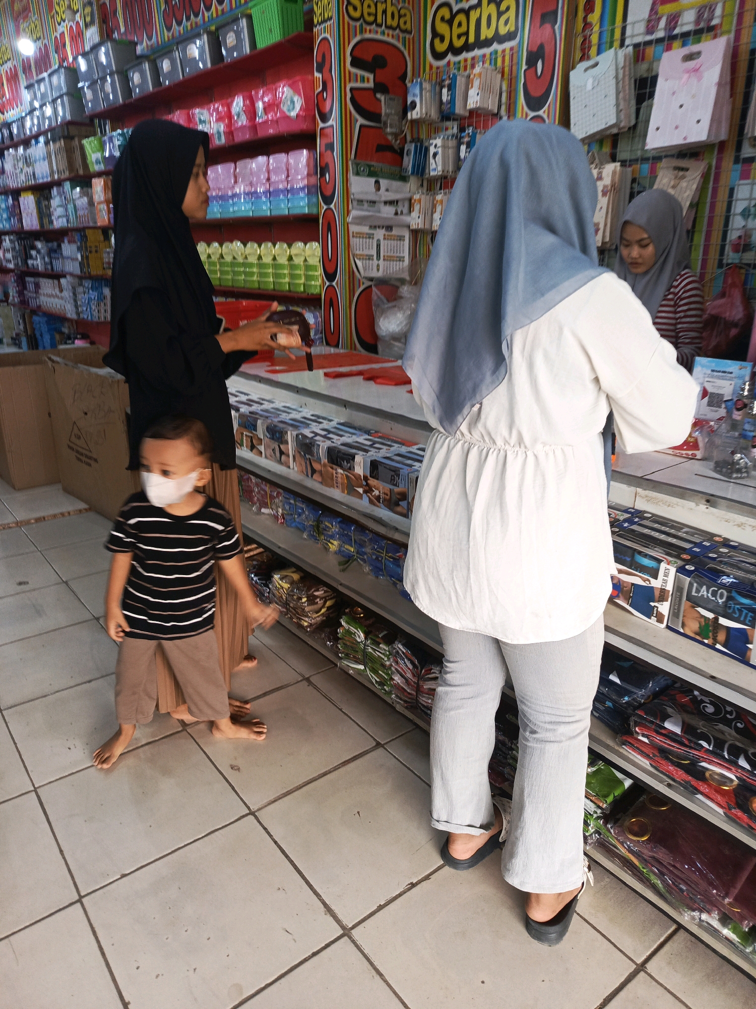 In a retail setting, a child wearing a striped t-shirt and light brown pants, with a white face mask, stands barefoot on a tiled floor. The child is positioned in front of two women. One woman, dressed in black, stands to the left, facing away from the viewer and holding a brown item. The other woman, wearing a light blue headscarf and a white top with light gray pants, is seen from behind, interacting with a display of items on shelves. Another woman, wearing a gray headscarf and a striped shirt, is partially visible in the background. The environment is a shop with shelves stocked with various products, including what appear to be clothing items and containers. The overall impression is a casual shopping scene.