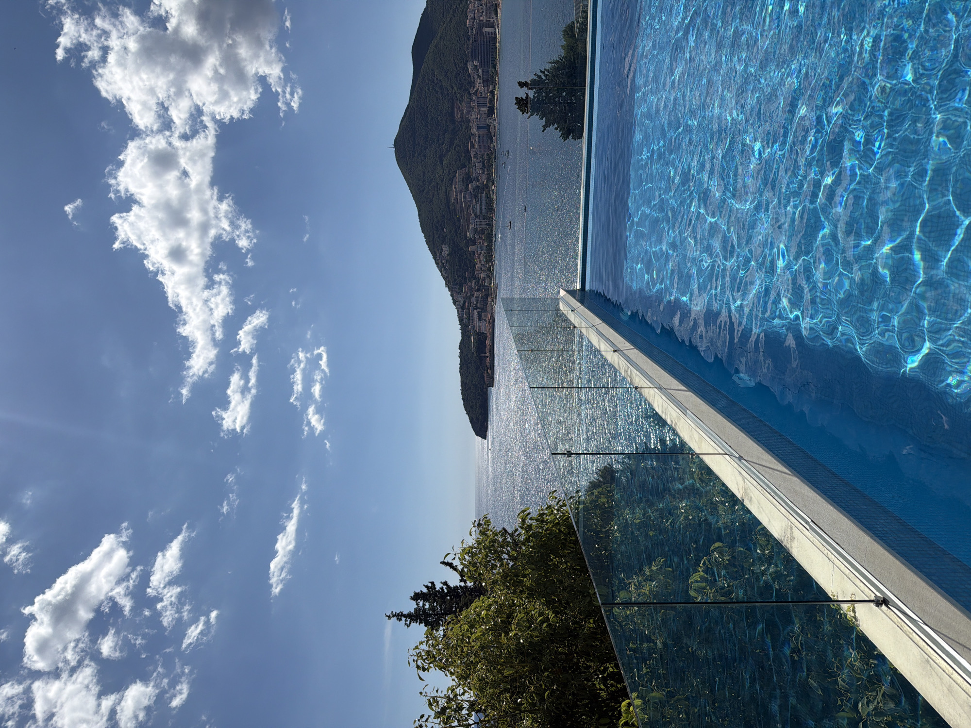 The image shows a swimming pool with crystal clear blue water, overlooking the Adriatic Sea and the city of Bečići, Montenegro. The pool has a modern glass fence. In the background, a lush green hill rises behind the city, which is dotted with buildings. The sea is sparkling under a bright, sunny sky with scattered white clouds. Several boats are visible on the water. Trees and foliage are present on the left side of the frame, adding to the natural beauty of the scene.