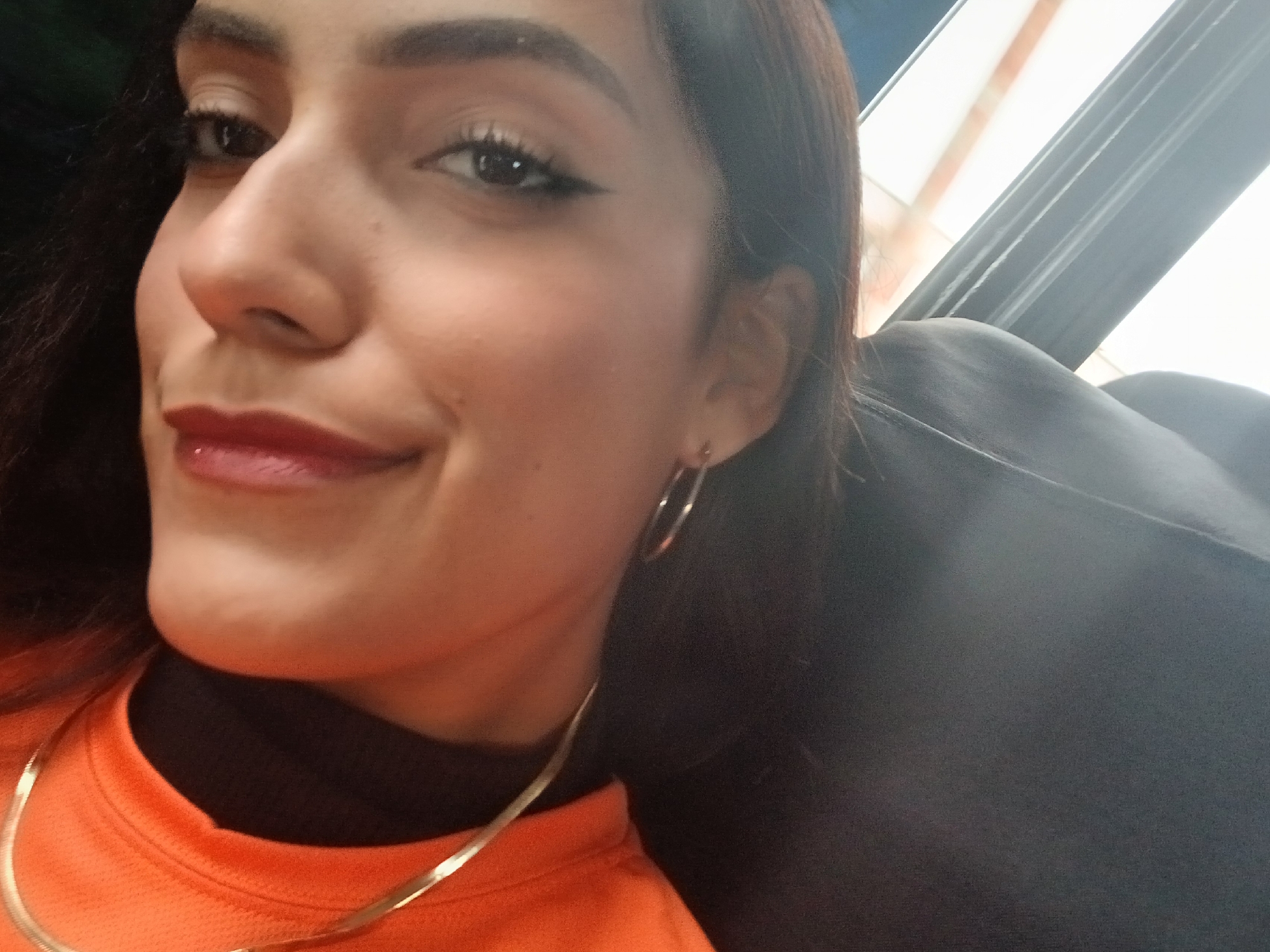 The image is a close-up, sideways selfie of a woman. She has dark hair, dark eyes with winged eyeliner, and red lipstick. She is wearing an orange top and large hoop earrings. The background appears to be indoors, with a dark surface and a window with blinds visible. There is no indication of Manzanillo, Venezuela, in the image.