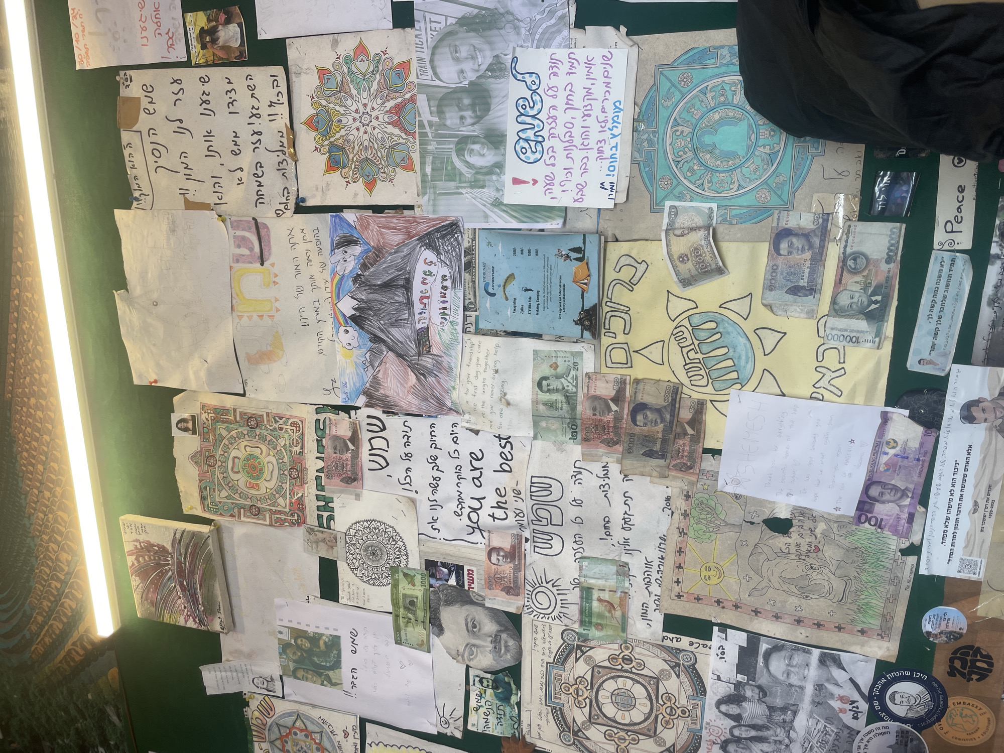 The image depicts a bulletin board or wall display in Bhagsu, India, covered with various papers, drawings, photographs, and currency. The items are pinned or taped to a dark green surface, with a portion of a light fixture visible at the top of the frame.
Prominent items include:
- Several intricate, mandala-like drawings in various colors, some featuring Buddhist or spiritual imagery.
- Handwritten notes and text in Hebrew, some accompanied by drawings or photographs.
- Several photographs, including group shots of people and individual portraits.
- Various denominations of foreign currency, appearing to be from different countries, are affixed to the display.
- A sketch of a rhinoceros.
- A drawing of a mountainous landscape with a rainbow and clouds.
- A printed flyer with adventure activity prices.
- A printed "TRAIN TICKET" with text in what appears to be English.
- A sticker with the word "EMBASSY" and an emblem.
- A sticker with text in Hebrew and the word "Peace".
The overall impression is a collection of personal mementos, messages, and artistic expressions, likely accumulated by travelers or residents in the Bhagsu area, which is known for its spiritual and backpacker culture. The density of items suggests a space where people share experiences and leave their mark.