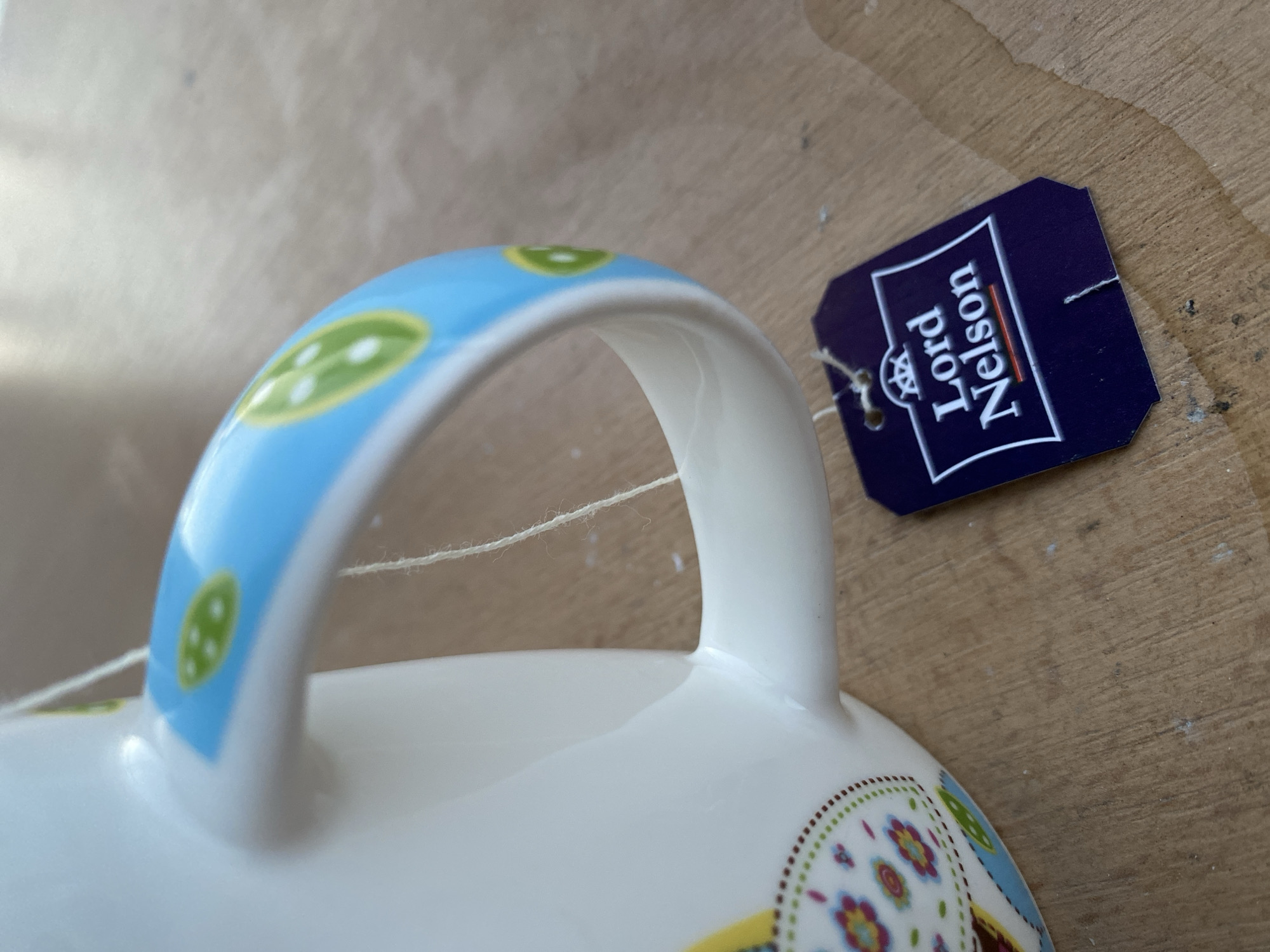 The image shows a close-up of a white mug with a blue handle decorated with green circles. A tea bag tag with "Lord Nelson" printed on it hangs from the mug by a white string. The mug and tea bag are resting on a wooden surface. The lighting suggests an indoor setting, possibly a kitchen or dining area. There are no people or specific landmarks visible to determine a location in Bílina, Czechia.