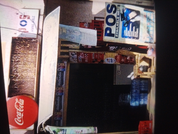 The scene depicts an outdoor commercial vending stall or shop frontage situated in Kaura Goje, Nigeria. The establishment offers Point-of-Sale (POS) services, evidenced by a prominent sign displaying "POS AVAILABLE HERE" and detailing services such as "CASH DEPOSIT," "WITHDRAWAL," "BILLS PAYMENT," and "TRANSFER." Another "POS AVAILABLE" sign is partially visible on the upper left.
The stall's merchandise includes stacked red Coca-Cola crates, some containing bottles, positioned on the right side. Several blue plastic crates filled with bottled water are visible below these crates. A red circular sign featuring the Coca-Cola logo rests on a wooden surface to the far left. Display units feature "Hollandia" branding. A dark, rectangular opening, likely a counter or service window, is centrally located, with a gray refrigerator or cooler visible behind it to the right. Various smaller packaged goods, including an item branded "Sweet Me" with "0000" below it, are arranged around the stall. A large, reflective, silver-colored object, possibly a metal sheet, is prominent on the left side of the frame, partially obscuring other elements. No individuals are present in the image, and no specific actions or interactions are depicted.