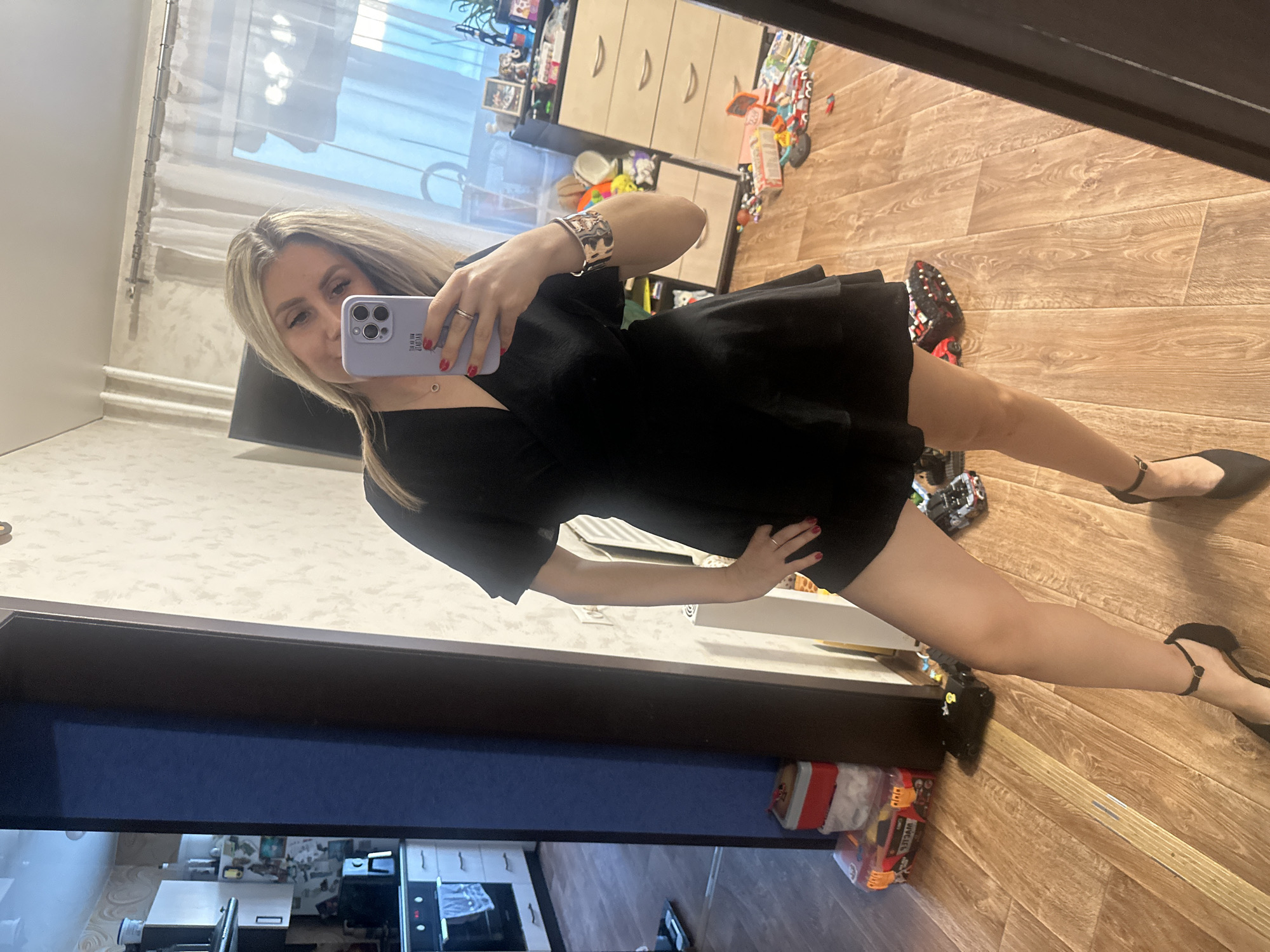 A woman is taking a mirror selfie indoors. She is wearing a black dress and black high-heeled shoes. She is also wearing a bracelet on her left arm and a ring on her left hand. She is holding a purple phone in her right hand, with which she is taking the photo. The background shows a room with wooden flooring, a wardrobe, and some toys. The lighting suggests it is daytime. The overall mood is casual and domestic. There is no visible text in the image. The location context is Novoaltaysk, Russia.