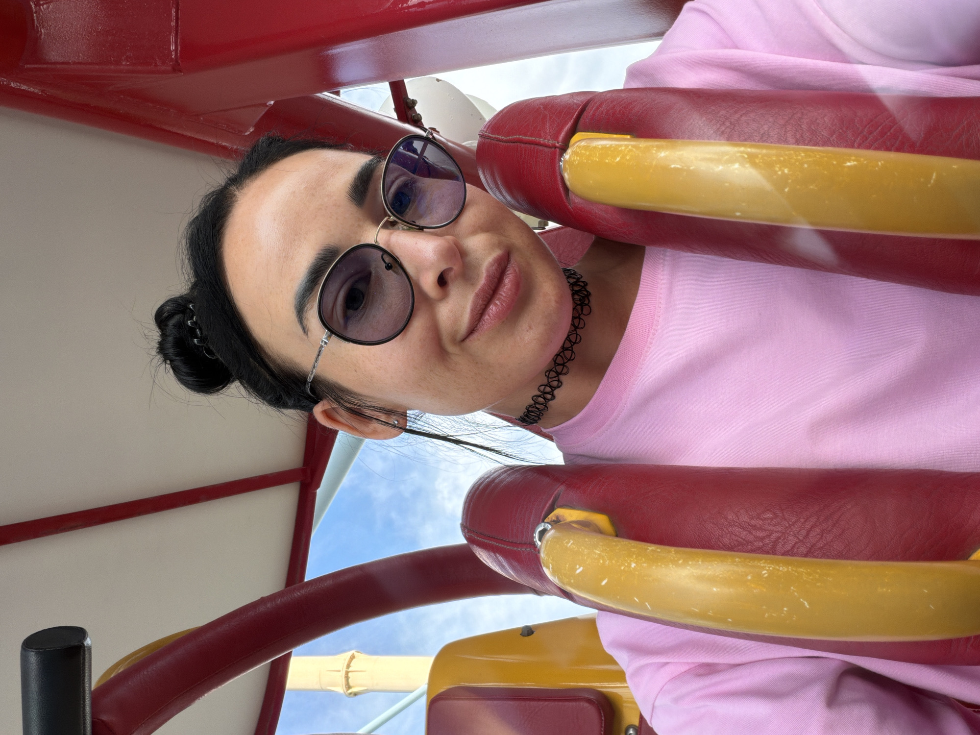 A woman is sitting in a red and yellow amusement park ride, possibly a roller coaster or a sling shot. She is wearing a pink t-shirt, sunglasses, and a black choker necklace. Her hair is pulled up in a bun. The sky is visible in the background, with some clouds. The image appears to be a selfie, taken by the woman herself. The location is not specified as Sochi, Russia.