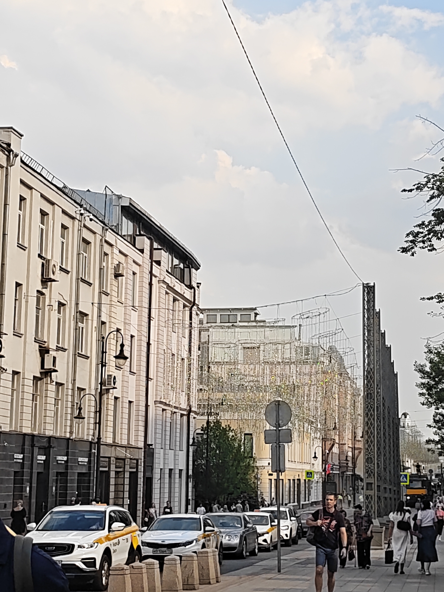 The image depicts a street scene in Moscow, Russia, likely on Tverskaya Street, known for its historical architecture and vibrant atmosphere. A procession of cars, including white taxis with yellow stripes, are parked and moving along the street. Pedestrians are seen walking on the sidewalk, some are dressed casually, suggesting a warm day. The buildings lining the street are of varying architectural styles, with some featuring ornate facades and balconies, while others have a more modern appearance. Decorative elements, possibly for an event or festival, are suspended above the street, adding to the festive ambiance. The sky is partly cloudy, with the sun casting a soft light on the scene. The overall impression is one of a bustling urban environment in Moscow, Russia, with a blend of historical and contemporary elements.