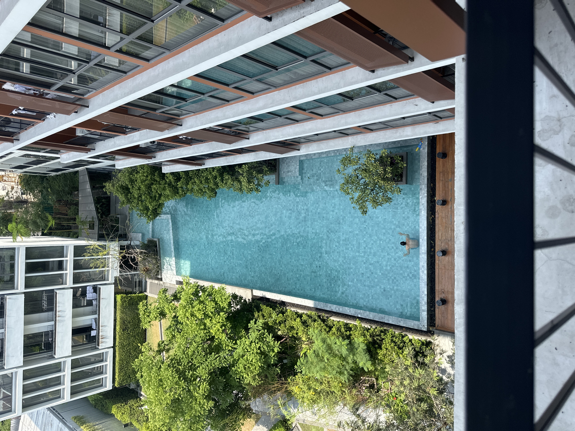 From an elevated perspective, the image depicts a rectangular swimming pool surrounded by buildings and greenery in an urban setting, presumably Bangkok, Thailand. One person is seen in the pool. The pool is adjacent to a wooden deck and surrounded by dense trees and shrubs. Two buildings flank either side of the pool area, and appear to be a residential or hotel development with multiple floors. Architectural details of the buildings suggest a modern design. The image is taken from a height where a portion of the viewing platform is visible.