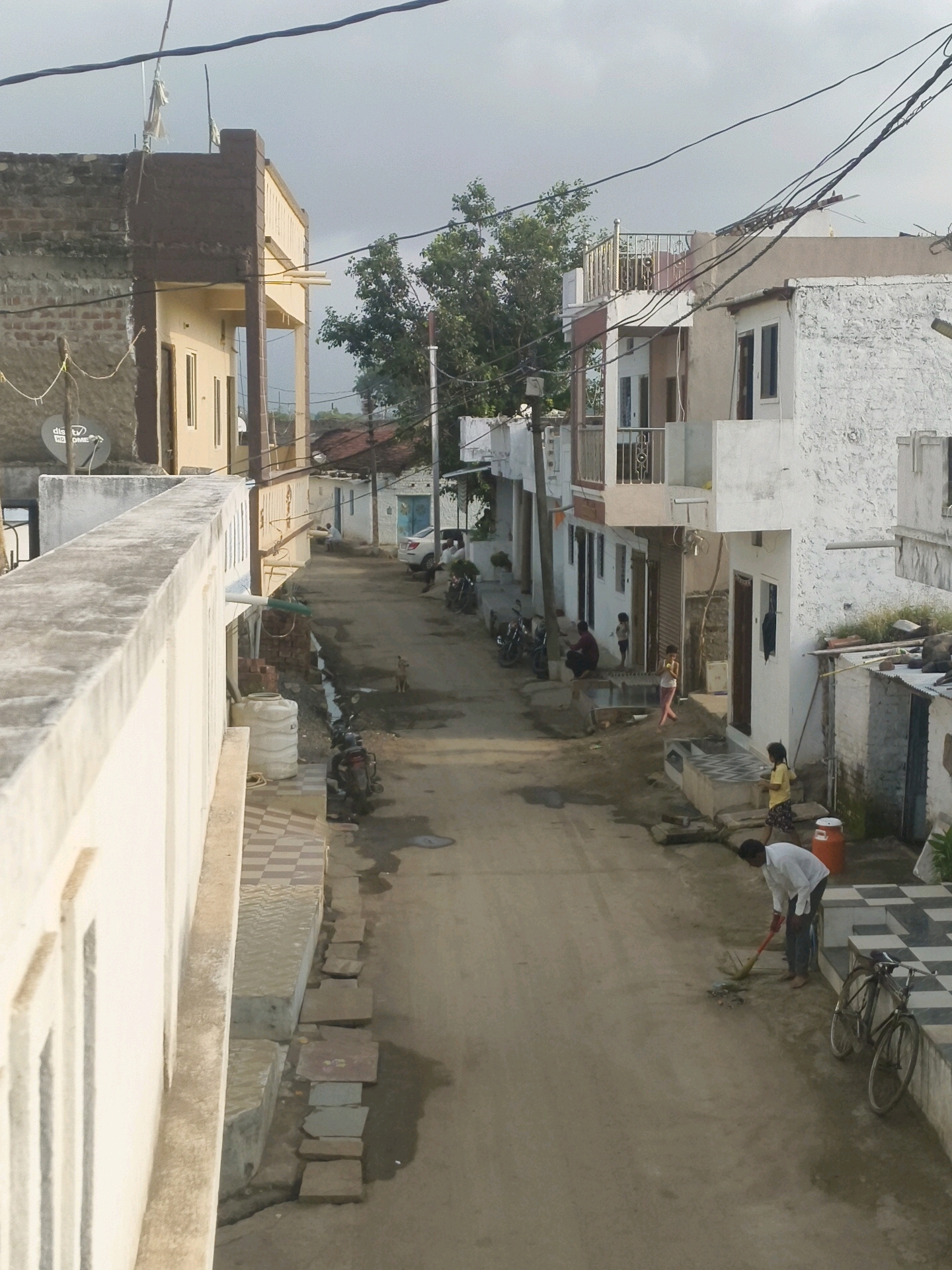 The image depicts a narrow, dusty street in Krodiya Khurd, India, lined with multi-story buildings. A man in a white shirt and dark pants is sweeping the street with a broom. A bicycle is parked nearby. Further down the street, several motorcycles are parked. A dog stands in the middle of the road. Children are visible playing and walking in front of the houses. The buildings are a mix of concrete and brick construction, with some featuring balconies and awnings. Overhead, power lines crisscross the sky, partially obscuring a cloudy sky. The overall impression is of a modest, lived-in urban environment.