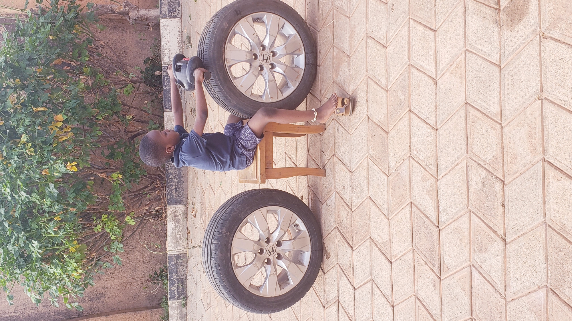 The image captures a young boy engaged in play outdoors in Narayi, Nigeria, during the daytime. The boy, who appears to be of elementary school age, has dark skin and short, dark hair. He is dressed in a dark blue short-sleeved polo shirt and patterned shorts, and is wearing light-colored sandals on his feet, with a shell anklet visible on his left ankle.
He is lying on his back on a small, light-colored wooden stool, positioned between two car wheels with tires. He is holding a gray and black object, possibly a toy or a tool like a car jack, above his head with both hands, seemingly mimicking lifting weights or performing a repair. One car wheel is located above his head, and the other is near his feet. Both wheels feature silver rims with a "H" logo, indicating they are Honda brand.
The setting is an outdoor paved area, characterized by light-colored interlocking paving stones arranged in a distinctive chevron or rhombus pattern. To the left of the boy and the wheels, there are lush green bushes and foliage, suggesting a garden or natural area alongside the paved ground. The overall lighting suggests it is a clear day. The boy appears focused and absorbed in his activity.
