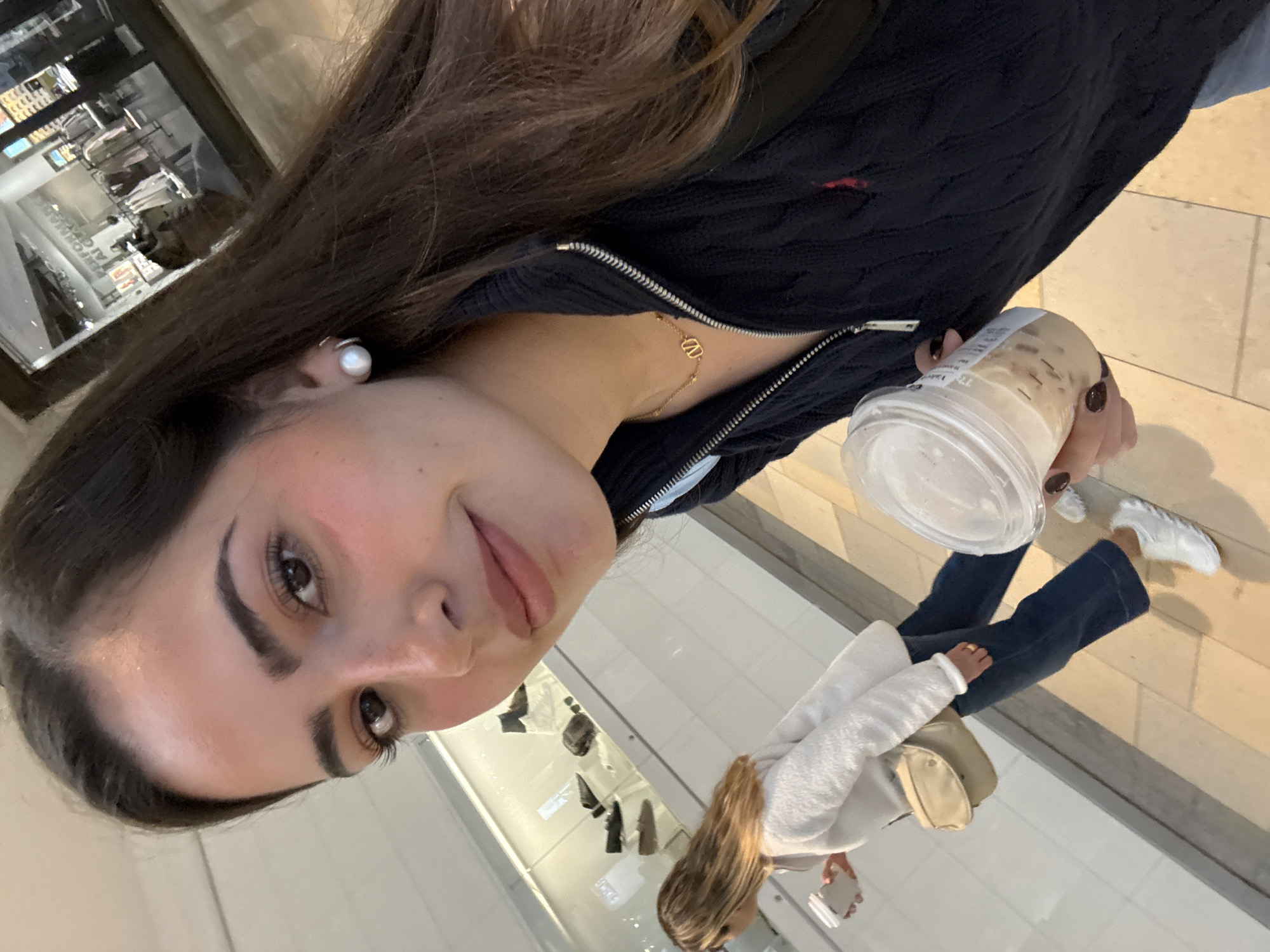 A woman holds an iced coffee in her right hand. She has dark hair and wears a black, zippered jacket with a small, red logo on the upper right side. A gold chain necklace with a "V" pendant is visible at her neck. She is wearing a pearl earring. The lower portion of the photo shows the reflection in a window. The reflection shows another person wearing a white sweater, dark jeans, and white sneakers. A beige purse hangs at their side. The floor is light-colored tile. The reflection in the window includes shoes for sale. It is difficult to say this image is San Antonio.