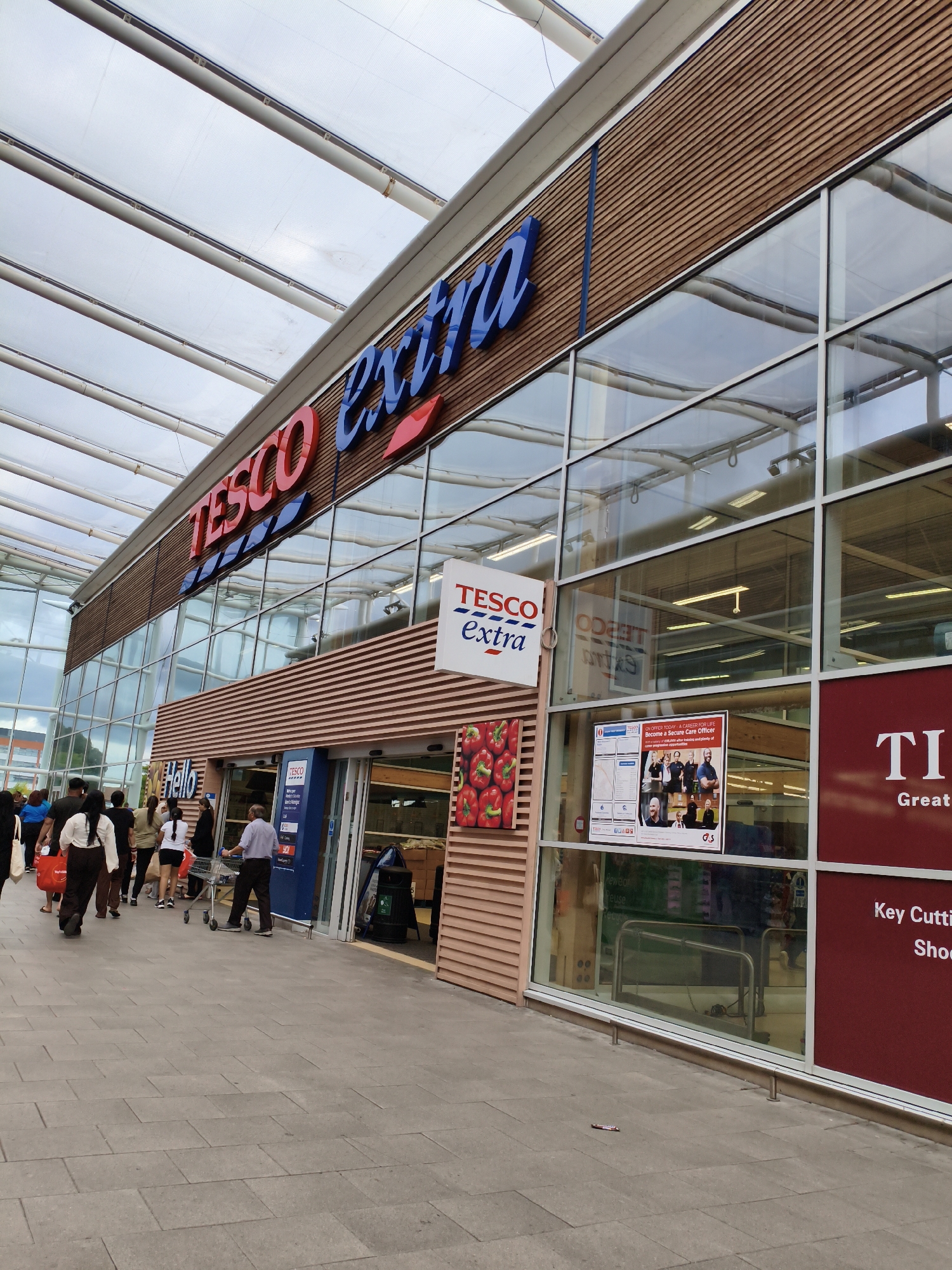 The image shows the exterior of a Tesco Extra supermarket in Sandwell, United Kingdom. The building has a modern design with a wooden-paneled facade and large glass windows. A large "TESCO Extra" sign is prominently displayed above the entrance. Several people are walking towards the entrance, some carrying shopping bags. One person is pushing a shopping cart. There are posters and advertisements displayed on the windows, including a recruitment advertisement for a "Secure Care Officer." The ground in front of the store is paved with grey tiles. The sky is overcast, suggesting a cloudy day.