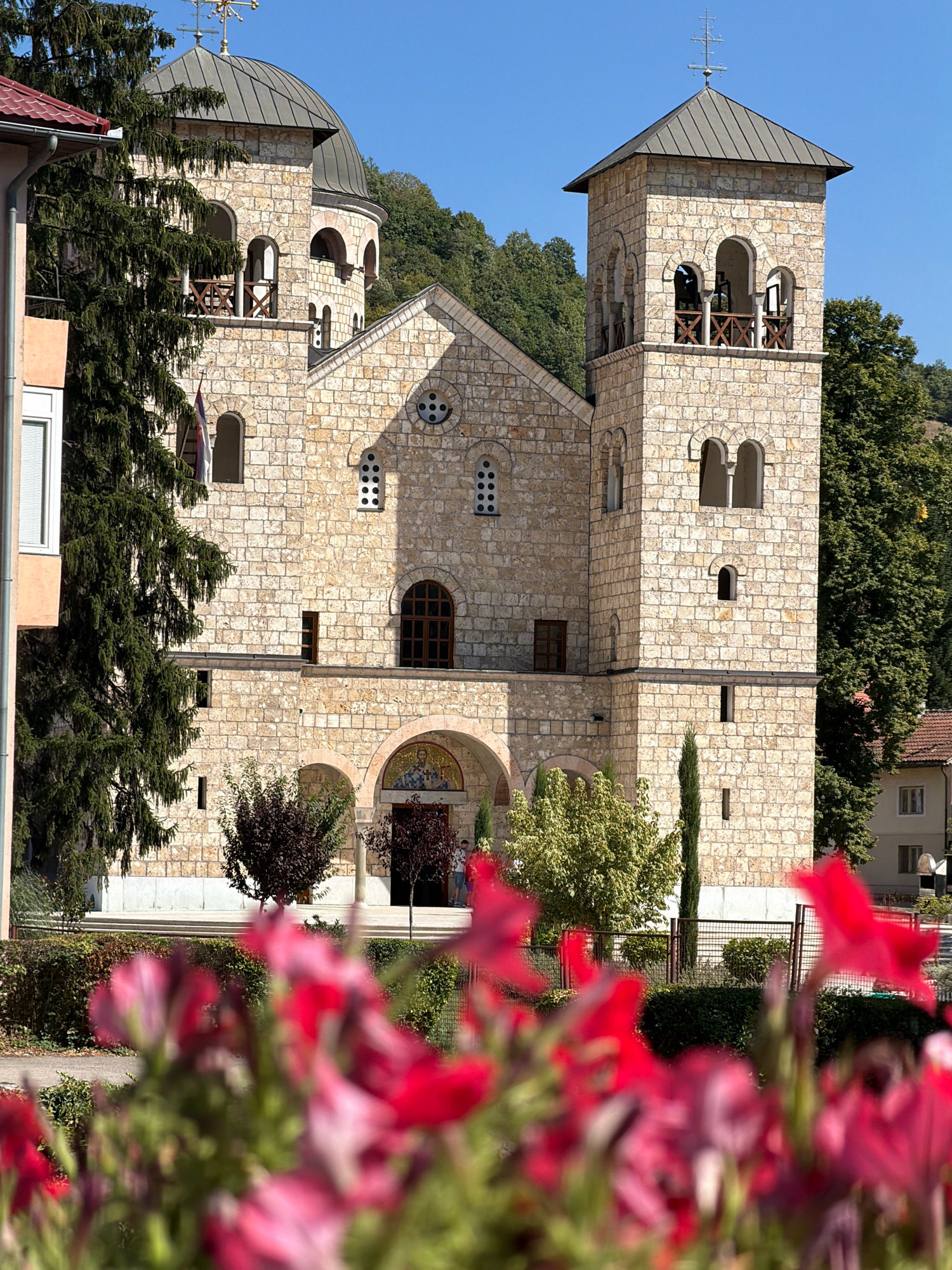 A stone church with two bell towers and a dome stands under a clear blue sky, with a blurred foreground of red and pink flowers. The church is built of light-colored stone blocks and features arched windows and doorways. A mosaic depicting a religious figure is visible above the main entrance. The church is surrounded by trees and some buildings are visible in the background. The location is the Serbian Orthodox Church of the Holy Trinity in Foča, Bosnia and Herzegovina. There are a few people visible near the entrance of the church.