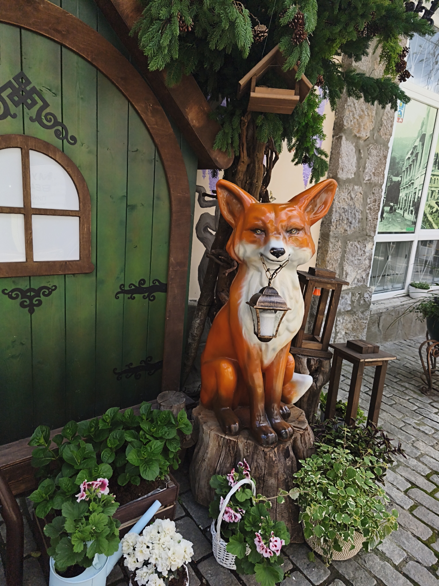 A colorful outdoor display in the city of Алупкинский городской совет, Ukraine, showcases a whimsical scene. A bright orange fox statue sits atop a tree stump, holding a lantern in its mouth. Behind the fox is a stylized green wooden door with a rounded top and small window. To the right, there's a tree with a birdhouse. A stone building is visible with a window. Assorted potted plants with green and pink flowers surround the fox statue and door. The ground is paved with cobblestones.