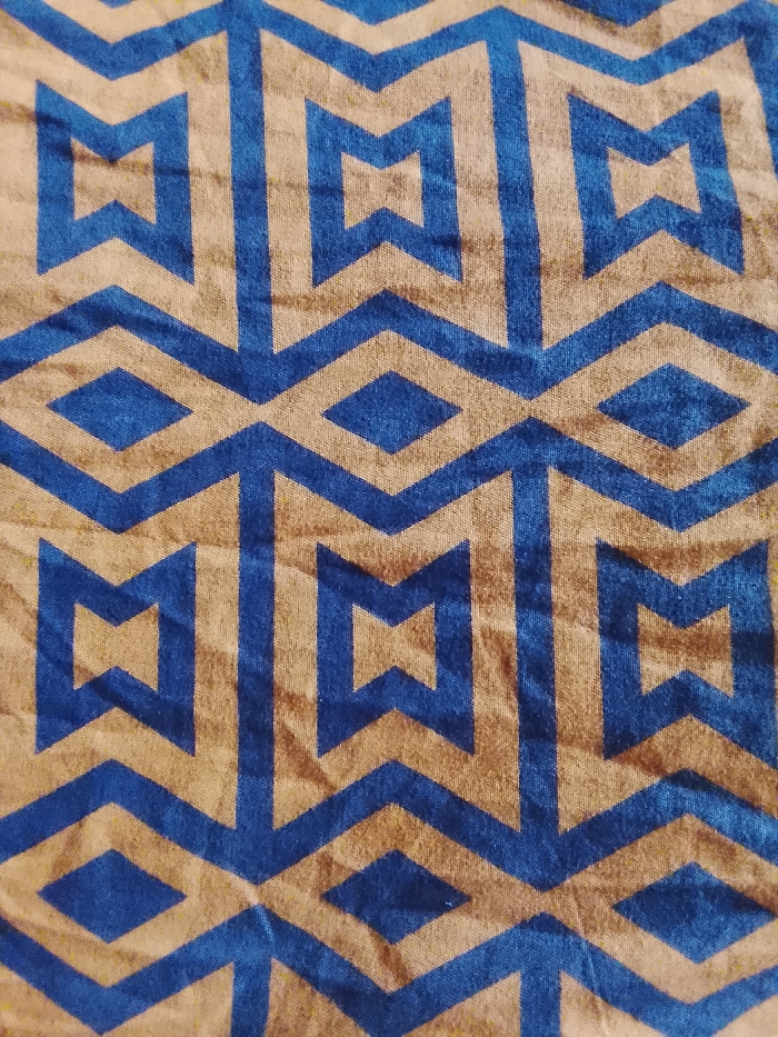 The image displays a close-up of a textile, located in Mgbidi, Nigeria. The fabric features a repeating geometric pattern composed of interlocking blue and tan elements. The blue forms include zig-zag lines, stacked diamonds, and stylized "T" or arrow-like shapes. These blue patterns are set against a tan background that similarly defines geometric outlines, creating a symmetrical visual rhythm across the surface. The textile exhibits minor creasing and a soft, slightly textured appearance. No individuals, objects other than the fabric, or specific environmental features are discernible within the frame. No actions or interactions are depicted.