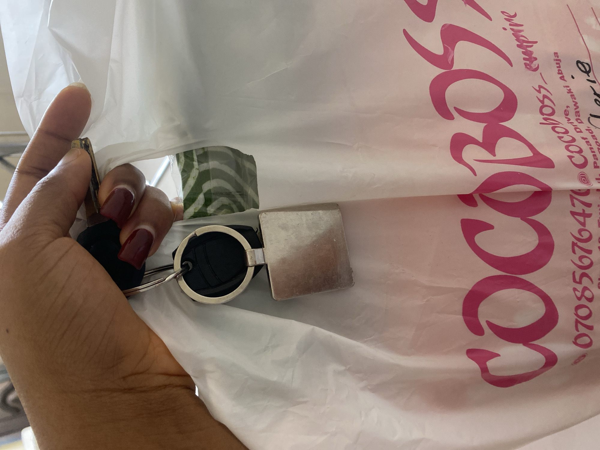 A person is holding a set of keys inside a white plastic shopping bag. The bag is printed with pink text that reads "CocoBoss_empire" followed by "Abuja", indicating the city. Below this, part of an address is visible: "4, Pan af, Road..." and a phone number "07085676476". The keys consist of a car key fob, a round, black fabric keychain, and a square metal tag.