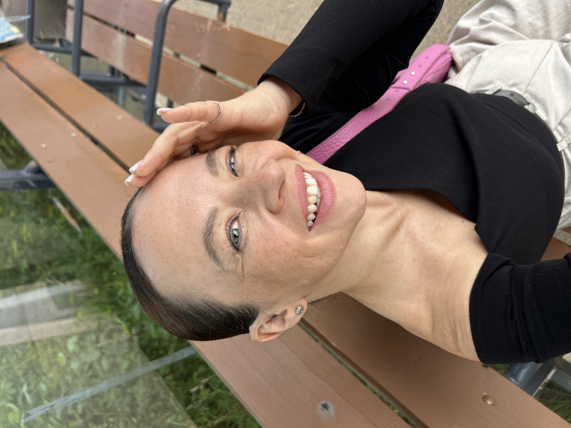 A woman with light skin and freckles is sitting on a wooden bench. She has her right hand on her forehead and is looking at the camera with a wide smile. She is wearing a black top with one shoulder exposed and a pink purse is visible on her left side. The bench appears to be outdoors, with greenery and a structure visible in the background. The scene suggests a casual outdoor setting, possibly a park or a recreational area. There are no indications that this location is in Chaikovsky, Russia.