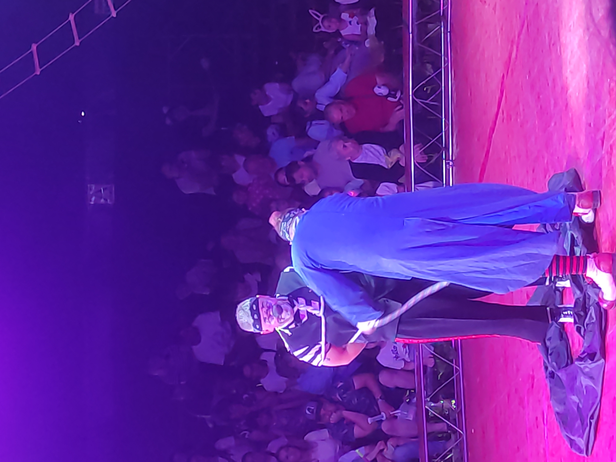In a circus tent, two clowns perform a routine on a pink stage. One clown, wearing a black and white striped vest, red and black striped pants, and oversized red and white shoes, has his arms bound by a rope. He wears a bandana, a white clown nose, and makeup. The second clown, dressed in a flowing blue robe and also wearing clown makeup and a red and white striped sock on one leg, appears to be interacting with the rope binding the first clown. The audience, seated in tiered rows illuminated by purple lighting, watches the performance. A ladder-like structure is visible in the upper right corner. The scene is taking place in the city of Mahilyow, Belarus.