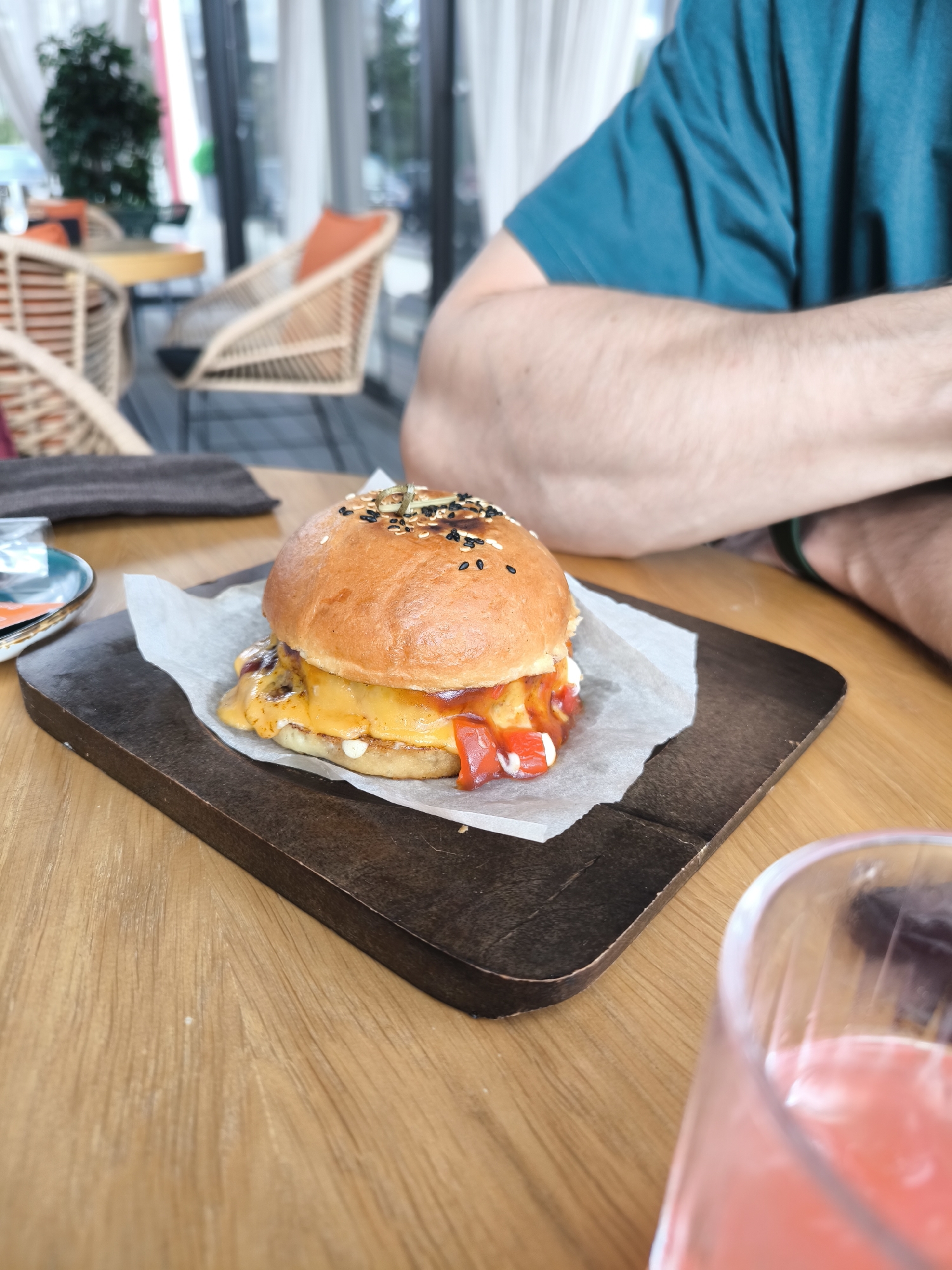 A close-up shot shows a burger served on a wooden board on a wooden table. The burger features a sesame seed bun, cheese, and red sauce. A glass with a pink beverage is also on the table, partially visible in the lower right corner. An individual wearing a teal shirt is partially visible in the background. The background includes furniture, potentially outdoor seating, and a glimpse of trees through a window. The location is Fryazino, Russia.