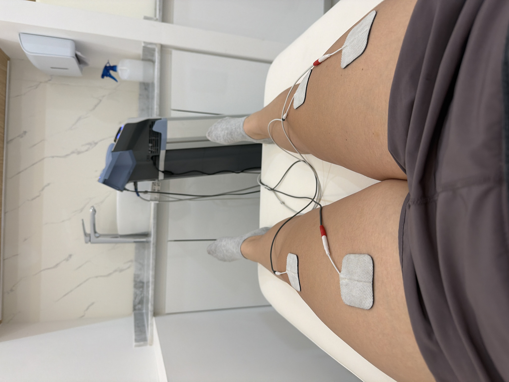 The image shows a person lying on a white examination table with electrodes attached to their upper thighs. The person is wearing gray socks and gray athletic shorts. Medical equipment is visible near the head of the table, including a device with a blue front panel, a sink, a soap dispenser, and a spray bottle. The background includes a white wall with a marble-like pattern. The location in San Bernardino Tlaxcalancingo, Mexico is not possible to verify with the current image.