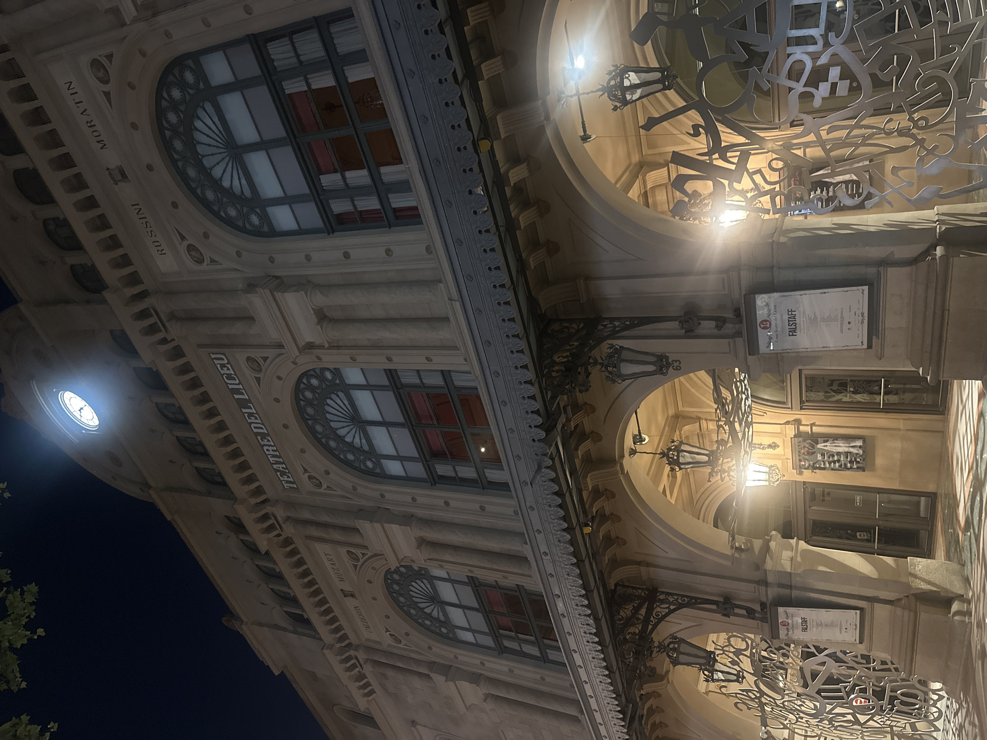 The image shows a facade of the Gran Teatre del Liceu in Barcelona, Spain, at night. The facade is made of light-colored stone and features ornate architectural details, including arched windows with decorative grilles and a patterned frieze. Above the entrance, the words "TEATRE DEL LICEU" are visible in large, decorative lettering.
Several names of composers and playwrights are inscribed on the facade, including "MORATIN," "ROSSINI," "MOZART," and "CALDERON." Two programs are displayed on the wall near an entrance. One program displays the word "FALSTAFF" in large lettering, along with the name "Gran Teatre del Liceu" and other text in Spanish.
The scene is illuminated by artificial lighting, including several ornate lamps and a bright spotlight shining on a clock face high on the building. The overall impression is one of elegance and grandeur, befitting a historic opera house. The time of day is clearly night, suggested by the dark sky and the artificial lighting.