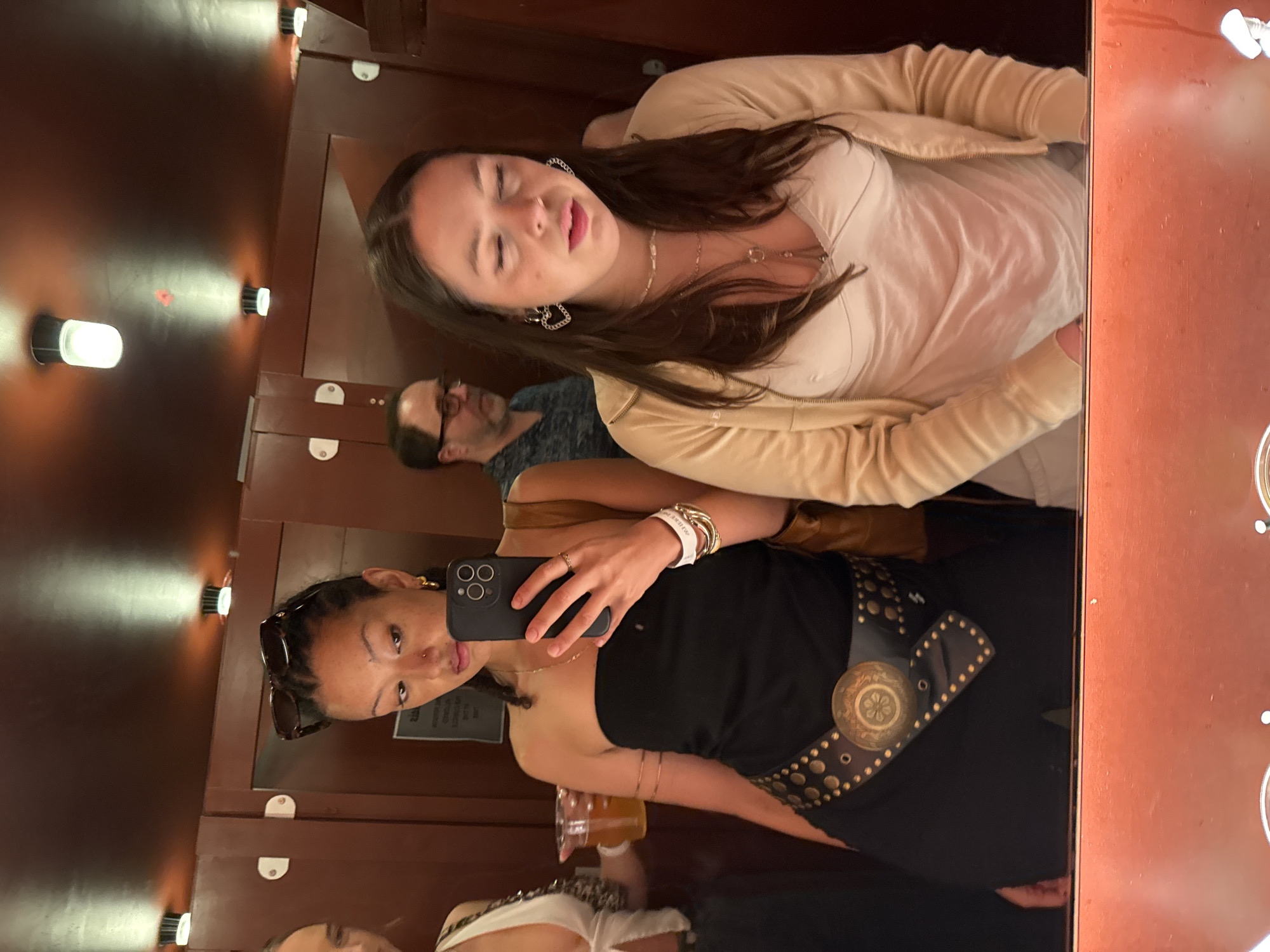 A group of people are taking a selfie in a narrow space with wooden walls and recessed lighting. One woman is holding the camera phone, wearing a black top and a studded belt. Another woman is standing next to her, wearing a light-colored top and a tan jacket. A man with glasses and a patterned shirt is visible in the background. The setting resembles a photo booth or a small elevator. The reflection suggests they might be in a public place with wood paneling, potentially in a bar or restaurant. Without additional context, pinpointing a specific location in London is not possible.