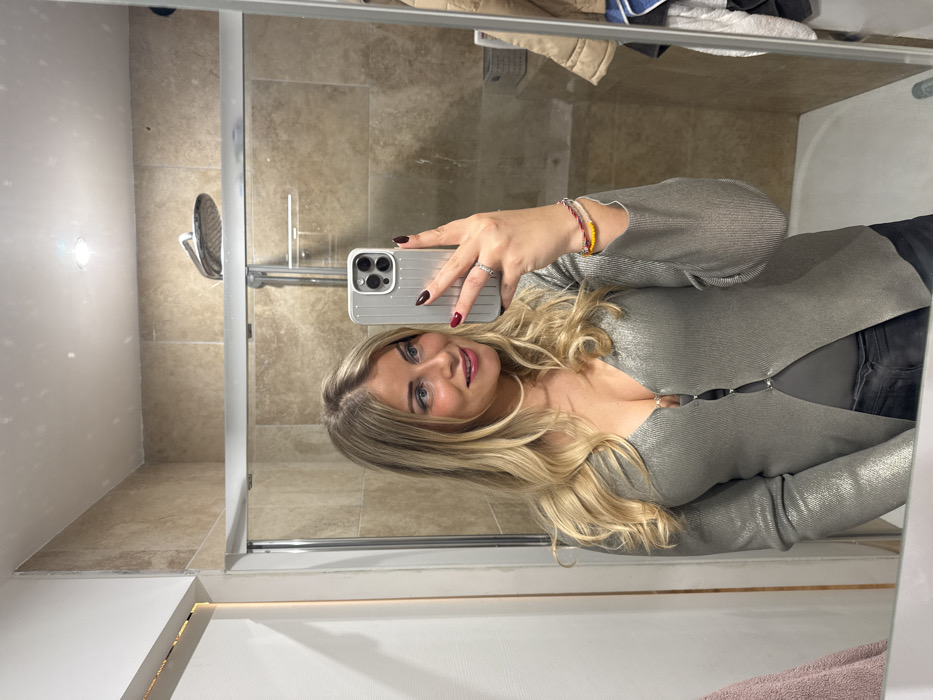 A blonde woman stands facing a mirror in a bathroom. She holds a phone, taking a selfie. She is wearing a silver top with three clasps down the middle and black pants. A shower is behind her in the reflection. The bathroom is decorated in neutral tones, with off-white walls and beige tiling in the shower area. A small light is reflected in the mirror.