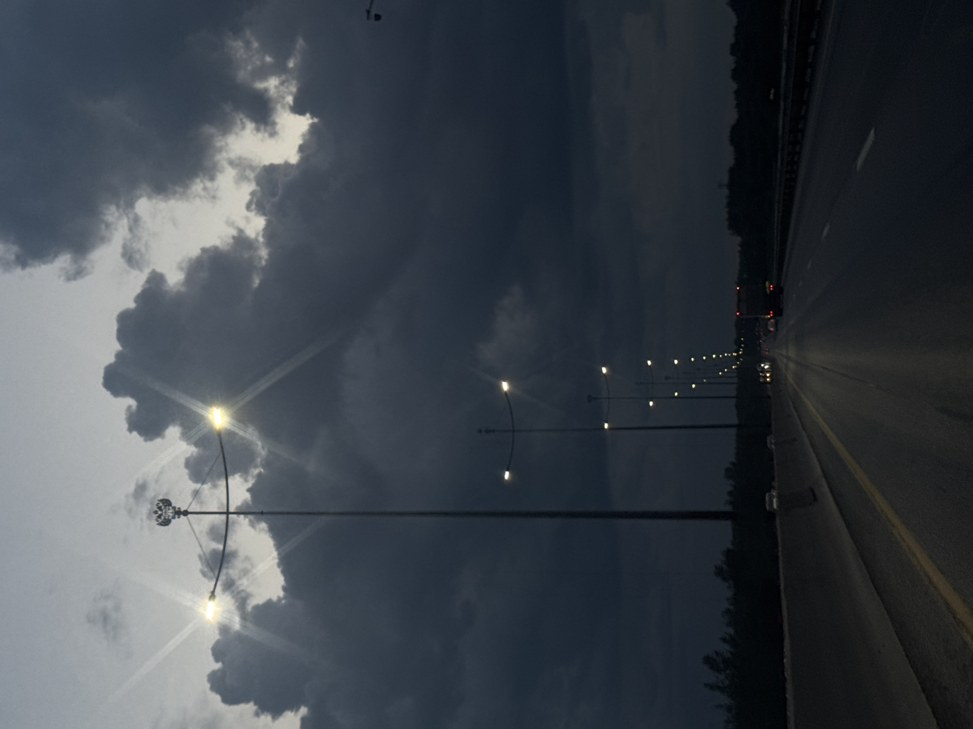 The image depicts a road in Kondratovo, Russia, at dusk or night. A multi-lane highway stretches into the distance, bordered by a concrete barrier on the left and trees or vegetation on the right. Streetlights, some with a double-headed eagle emblem on the tallest pole, illuminate the road. The sky is dominated by dark, heavy clouds, with hints of lighter sky visible through breaks. A few vehicles are present on the road, indicated by their headlights and taillights. The overall atmosphere is moody and dark, suggesting a potentially overcast or stormy evening.