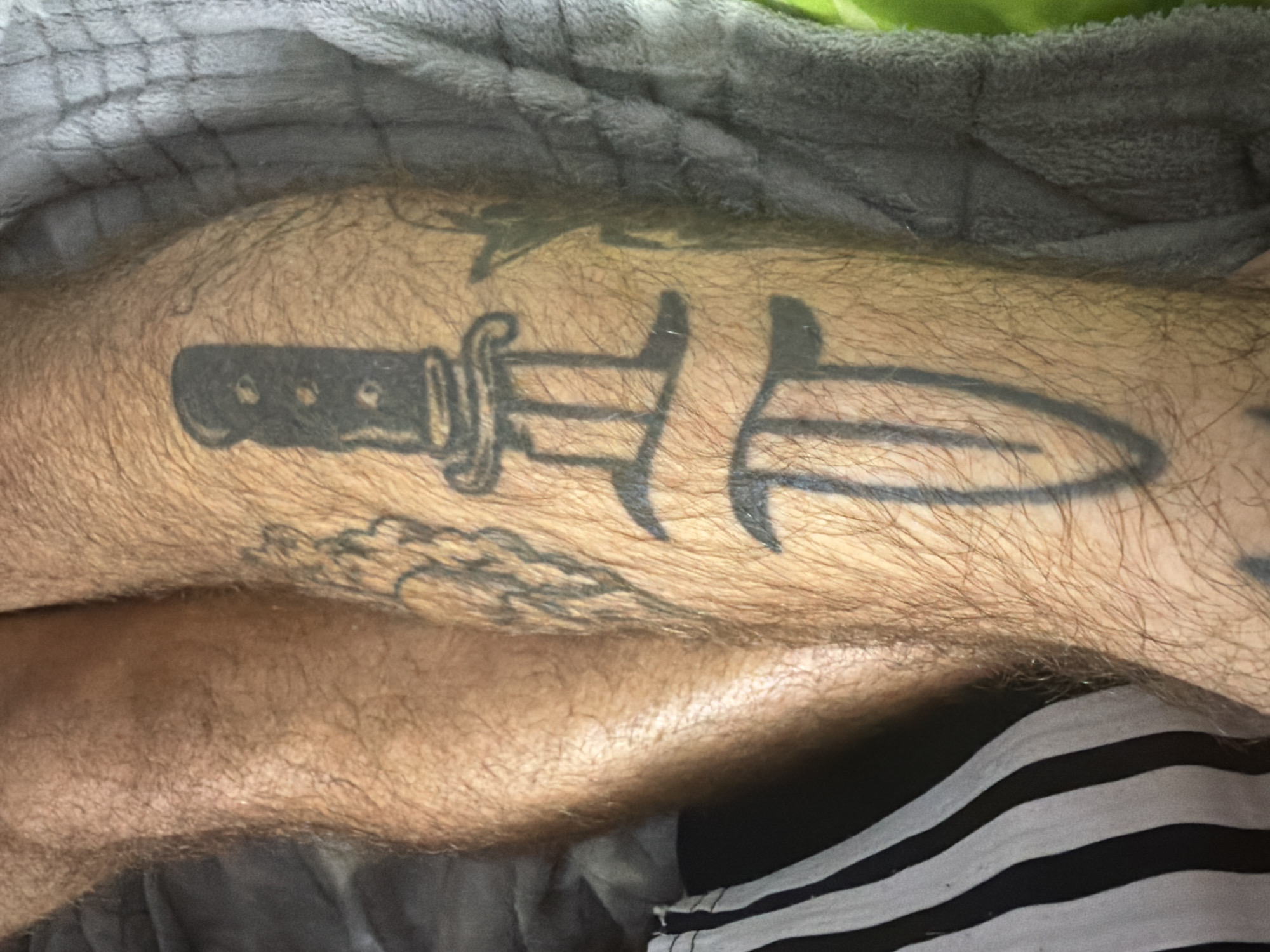The image shows a close-up of a person's leg with tattoos. The tattoos appear to be black ink on skin. One tattoo is a stylized dagger or sword, depicted vertically. Another tattoo, partially visible on the left, seems to be a more intricate design with lines and shading. The leg is covered in hair and is resting on a grey, textured fabric, possibly a blanket. On the lower left side of the image, a portion of black and white striped fabric is visible, possibly clothing. The background is a blur of grey and green. The location in Dorsten, Germany, cannot be determined from the visual information provided in the image.