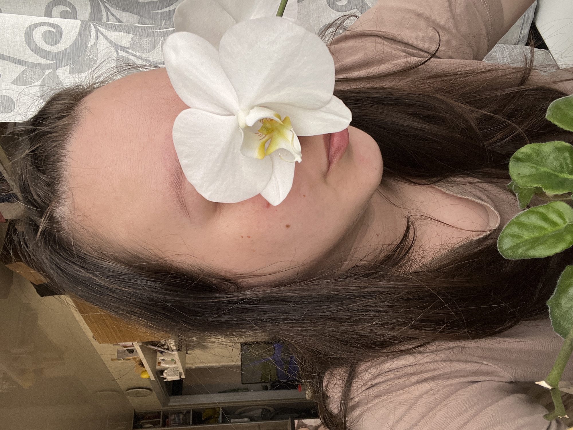 A person lies on their back. A white orchid flower is placed on the person's forehead, obscuring one eye. The person has dark hair and is wearing a light brown shirt. A plant with green leaves is visible to the right. The background shows a glimpse of a cluttered room.