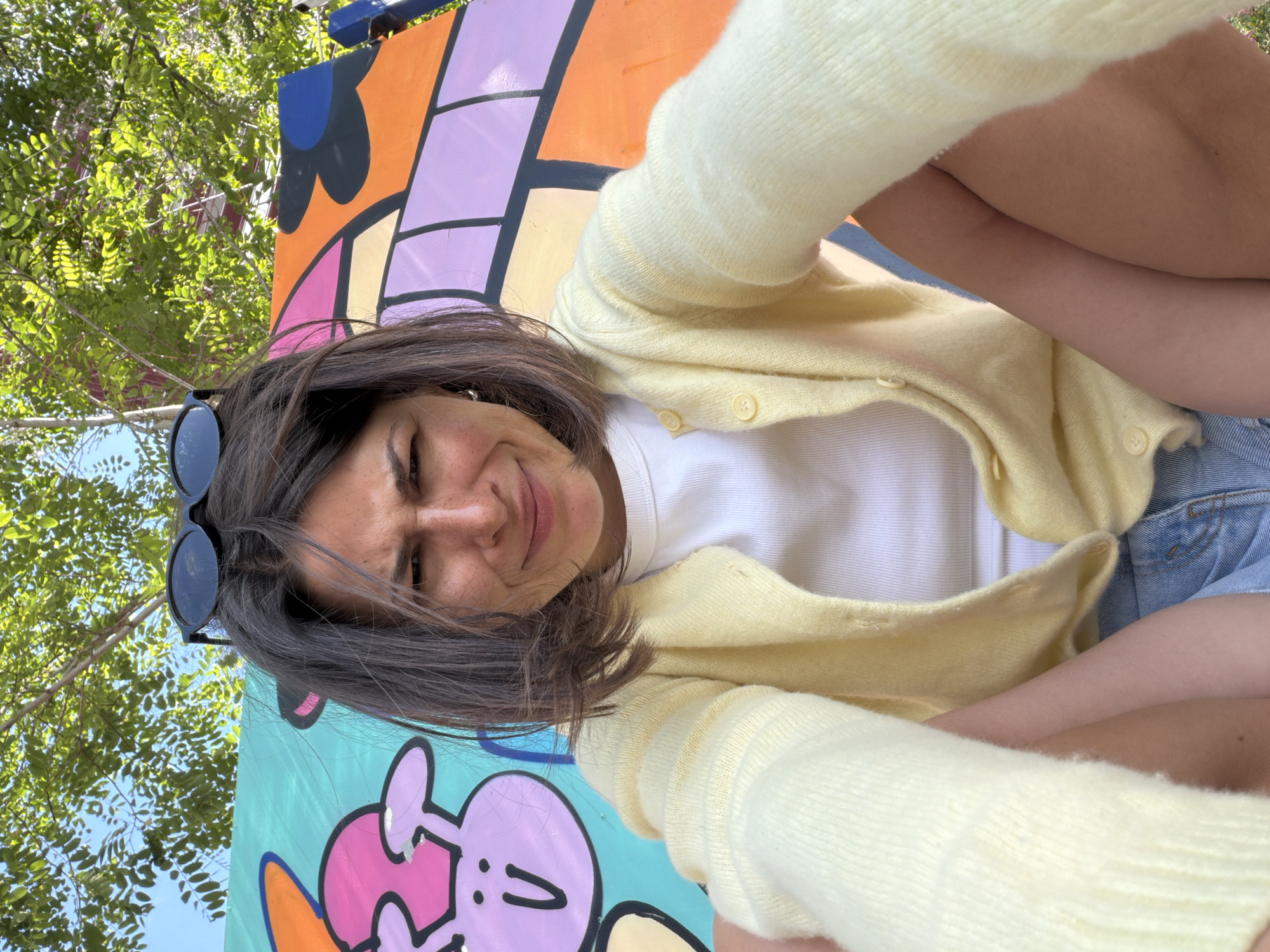 A woman in a yellow cardigan and white shirt poses for a selfie in front of a colorful, graffiti-covered structure. Sunglasses are perched on her head. Blue denim shorts can be seen. The location appears to be outdoors, possibly Rafailovići, Montenegro. Trees can be seen in the upper left of the image.