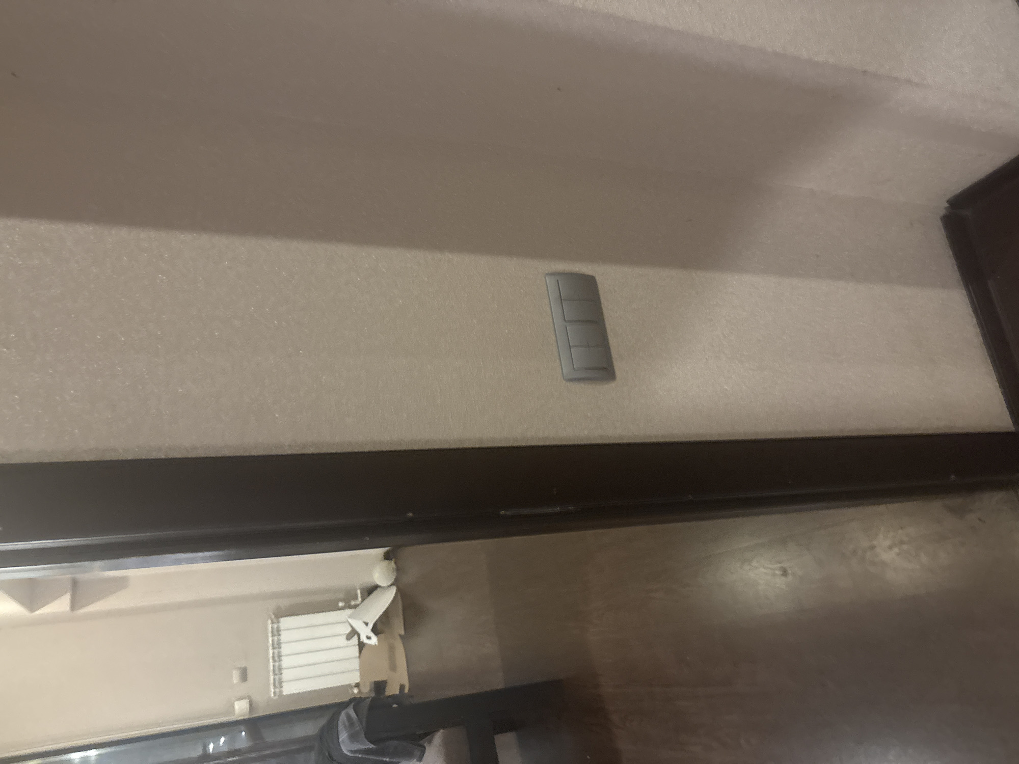 The image shows an interior scene, possibly a hallway or doorway. In the foreground, a dark brown door frame is visible on the right side. A light-colored wall with a textured finish is to the right of the door frame. Mounted on this wall is a gray light switch plate with three toggle buttons.
To the left of the door frame, a portion of a room is visible through an open doorway. This area has a dark wooden floor. In the background of this room, a white radiator is mounted on a light-colored wall. There are also some cardboard boxes and a white object on the floor near the radiator. The overall lighting is dim. There are no people visible in the image. The location is not specified in the image, nor can it be inferred from the visual content.