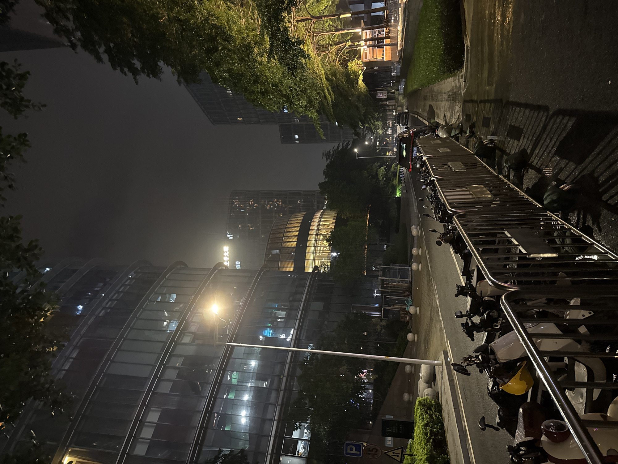 This image depicts a nighttime scene in Haizhu District, China. The focus is on a street lined with modern glass-fronted buildings, illuminated by streetlights and the lights from within the offices. A row of motorcycles is parked along a metal barrier on the right side of the frame, with a yellow helmet visible among them. The street appears wet, suggesting recent rain or it being a damp evening.
Trees with dense foliage are visible, casting shadows on the wet pavement. The atmosphere is somewhat moody due to the darkness and the rain. The buildings have distinct architectural features, with curved facades and numerous windows. The time of day is clearly night, indicated by the artificial lighting and the dark sky. There is no discernible activity or event taking place, other than the presence of parked vehicles and the illuminated buildings, suggesting a quiet urban environment after dark.
There is some faint text visible on signs in the distance, but it is not legible enough to be read. The overall impression is one of a contemporary urban landscape at night, possibly after a rainfall, with a focus on the juxtaposition of modern architecture and the parked motorcycles.