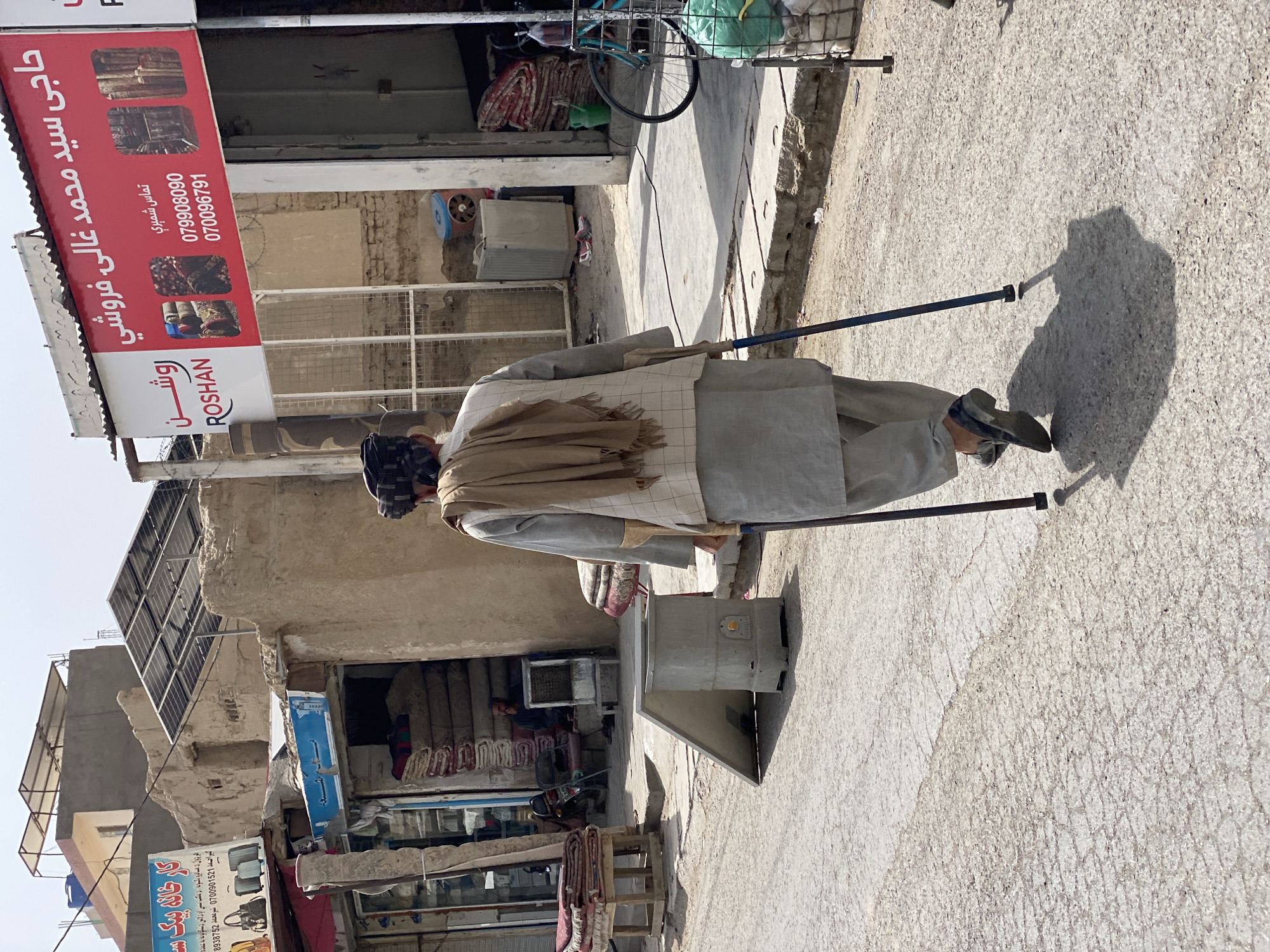 A man with a disability, using two crutches, walks away from the camera down a paved street in Kandahar, Afghanistan. He is wearing traditional clothing: a light-colored tunic and pants, a checkered vest, and a brown scarf draped over his shoulders. The street appears to be lined with shops and buildings constructed from mud brick. A blue and red sign for "ROSHAN" is prominently displayed on a building to the right, featuring Arabic and English text. Stacks of rugs are visible outside some of the shops. A bicycle is parked to the right of the Roshan sign. The environment suggests a bustling marketplace in the city.