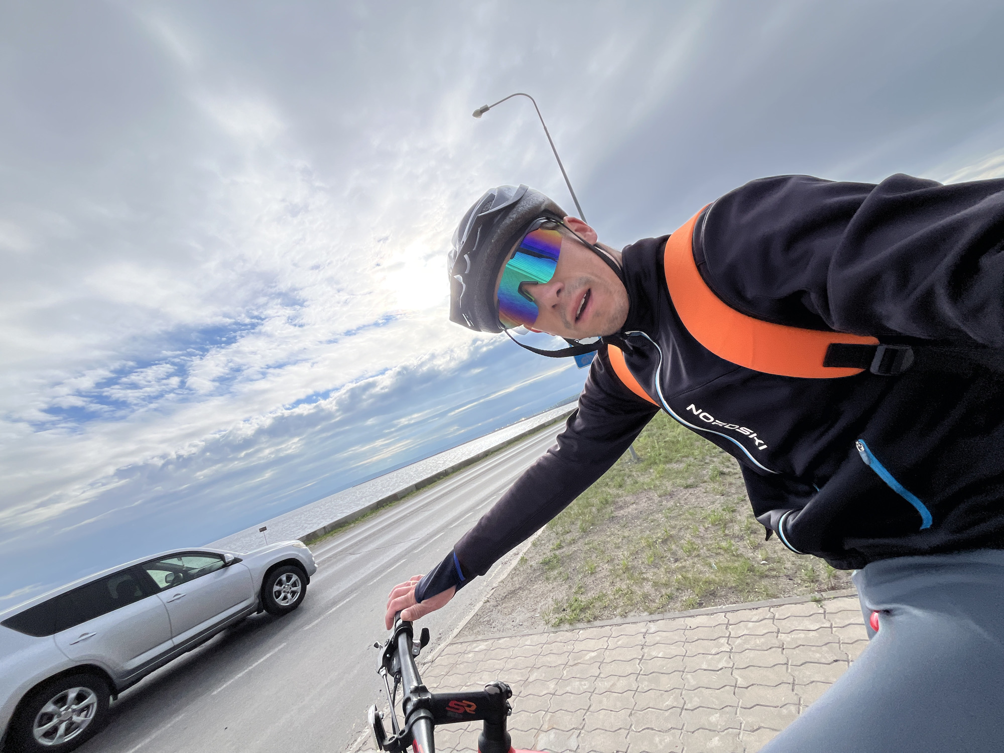 This is a first-person perspective selfie of a man cycling in Chaikovsky, Russia. The man is wearing a black cycling helmet, mirrored cycling glasses with rainbow lenses, a black long-sleeved cycling jacket with an orange high-visibility vest over it, and gray cycling shorts. He is holding the handlebars of his red bicycle. To the left of the frame, a silver SUV is driving on the adjacent road. The road runs alongside a body of water, likely the Kama River, with a cloudy sky overhead and a streetlamp visible in the background. The pavement is made of interlocking grey concrete blocks.