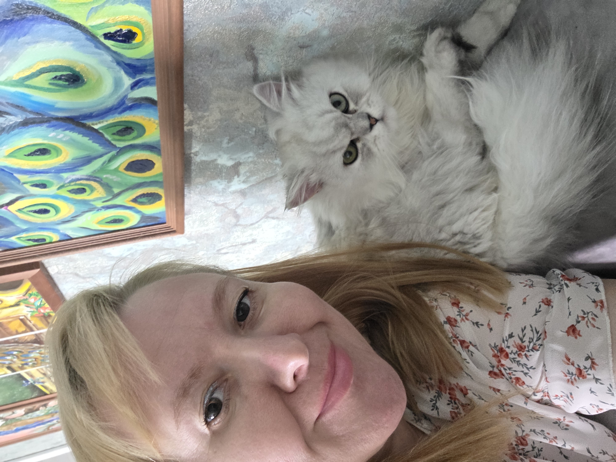 The image is a close-up selfie taken indoors, likely in an apartment. A woman with blonde hair and fair skin is partially visible on the left side of the frame, looking towards the camera with a slight smile. She is wearing a light-colored blouse with a floral pattern. To her right, a fluffy white Persian cat with green eyes is looking directly at the camera with a somewhat displeased expression. The background features a textured wall and a framed painting of peacock feathers in vibrant blue, green, and yellow colors. The overall impression is a casual, candid moment captured between a woman and her pet. The location of Novocheboksarsk, Russia, is not discernible from the image content.