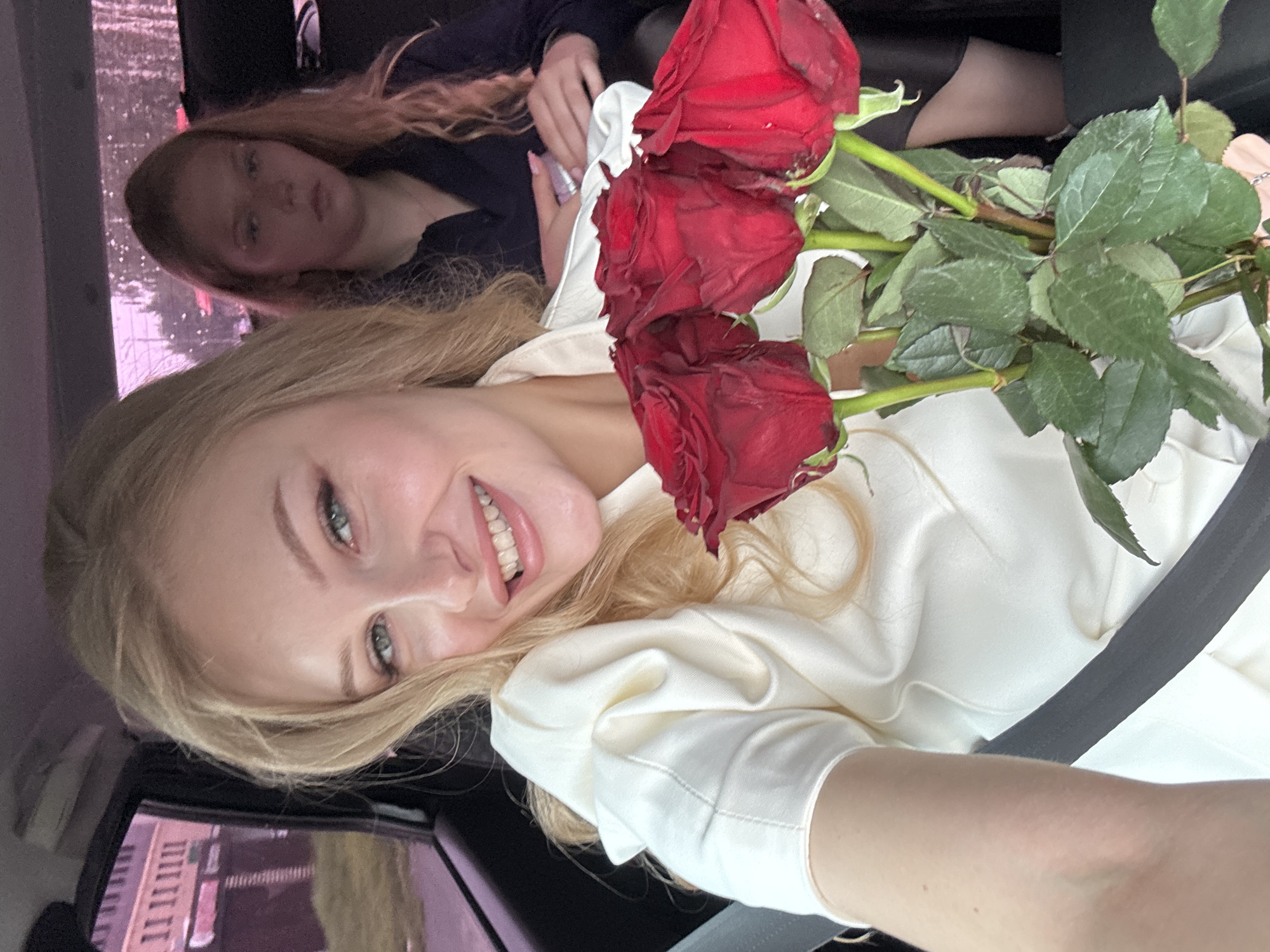 Two women are inside a car, with one woman in the foreground holding a bouquet of red roses. The woman in the foreground, with blonde hair and blue eyes, is smiling and looking at the camera. She is wearing a white blouse. The woman in the background has blonde hair and appears to be looking down. She is wearing a dark blue top. The car appears to be moving, as suggested by the blurred background outside the windows. The scene is consistent with a car ride within the city of Perm, Russia.