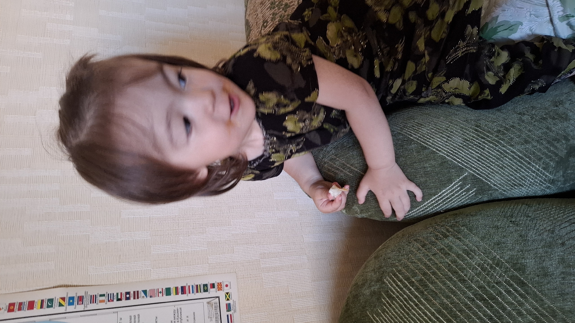 A young girl with brown hair and fair skin is leaning on a green upholstered sofa. She is wearing a dark dress with a green floral pattern. Her right hand is holding a small piece of food, and her mouth is slightly open. The background features a textured wallpaper and a poster on the left wall with flags of various countries. The image appears to be taken indoors in a residential setting. The poster on the wall is part of an educational display related to geography or international affairs, potentially in a home or classroom environment in Mednoe-Vlasovo, Russia.