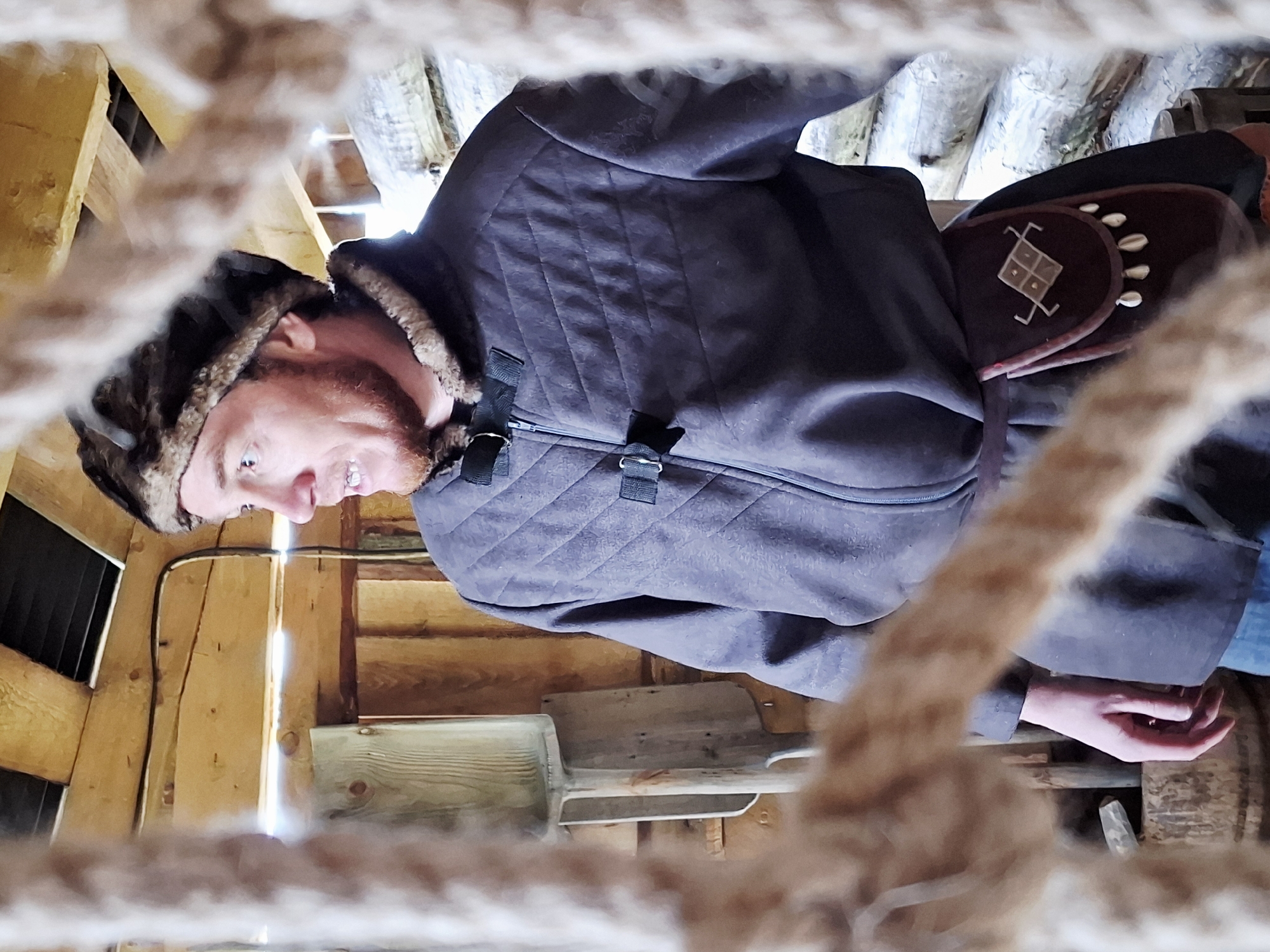 A medium shot photograph depicts a man inside a wood structure, taken from a low angle through a rope net. The man, wearing a fur-lined hat and a quilted jacket, looks to the left with a slight smile. A decorative pouch hangs at his side. The background features wooden walls and a spade leaning against the wall. The image does not provide enough information to determine if the location is in the city of Адам, Russia.