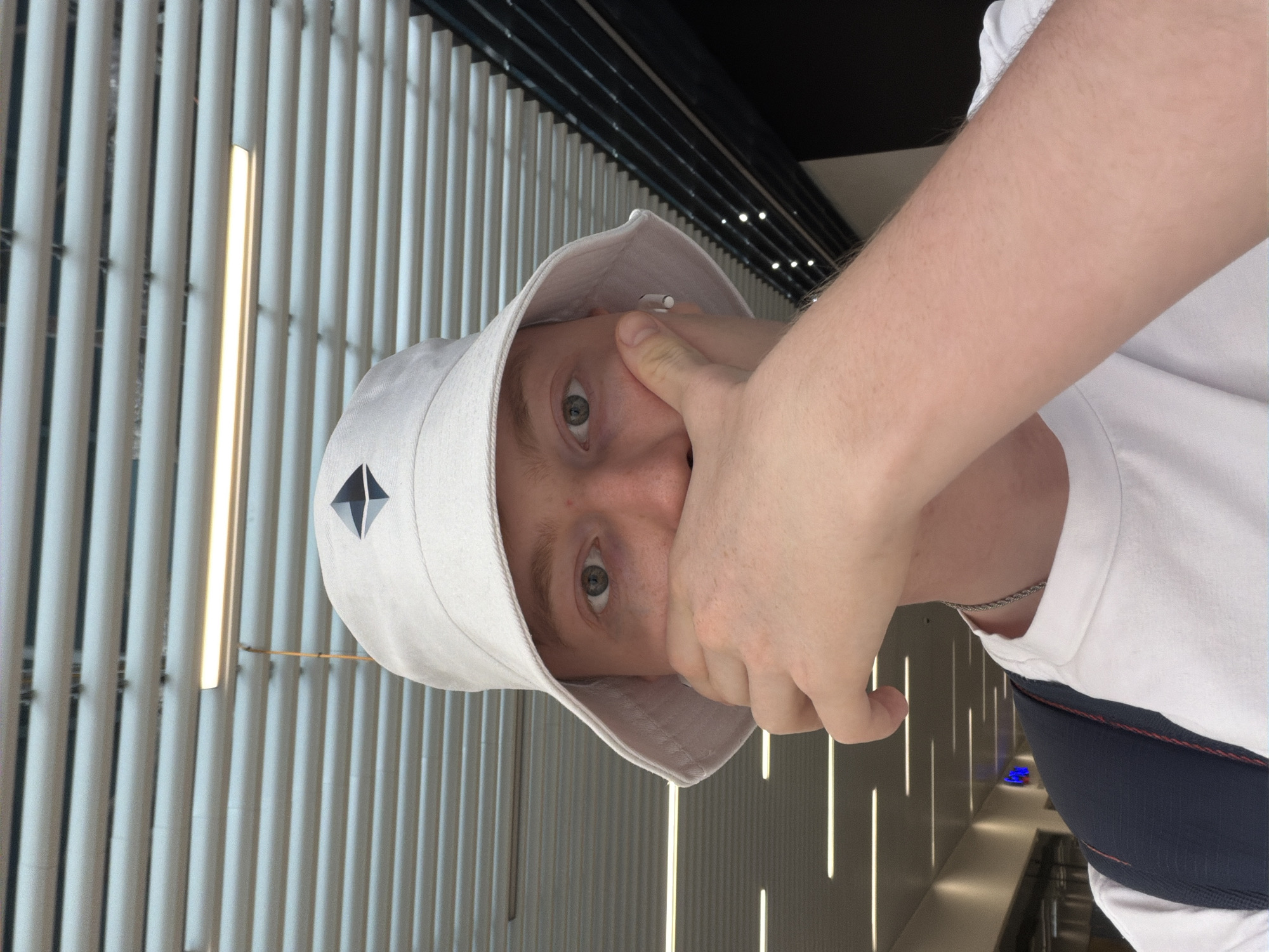 A close-up, selfie-style image shows a young person wearing a white bucket hat with a black diamond-shaped emblem. The person's right hand is partially covering their mouth. They are wearing a white t-shirt and a silver chain necklace. A dark blue backpack strap is visible over their shoulder. The background appears to be an indoor public space with a ceiling of horizontal white blinds and strip lighting. The environment suggests a modern building, possibly a shopping mall or an airport terminal. There is no indication of the location being Kyiv, Ukraine.