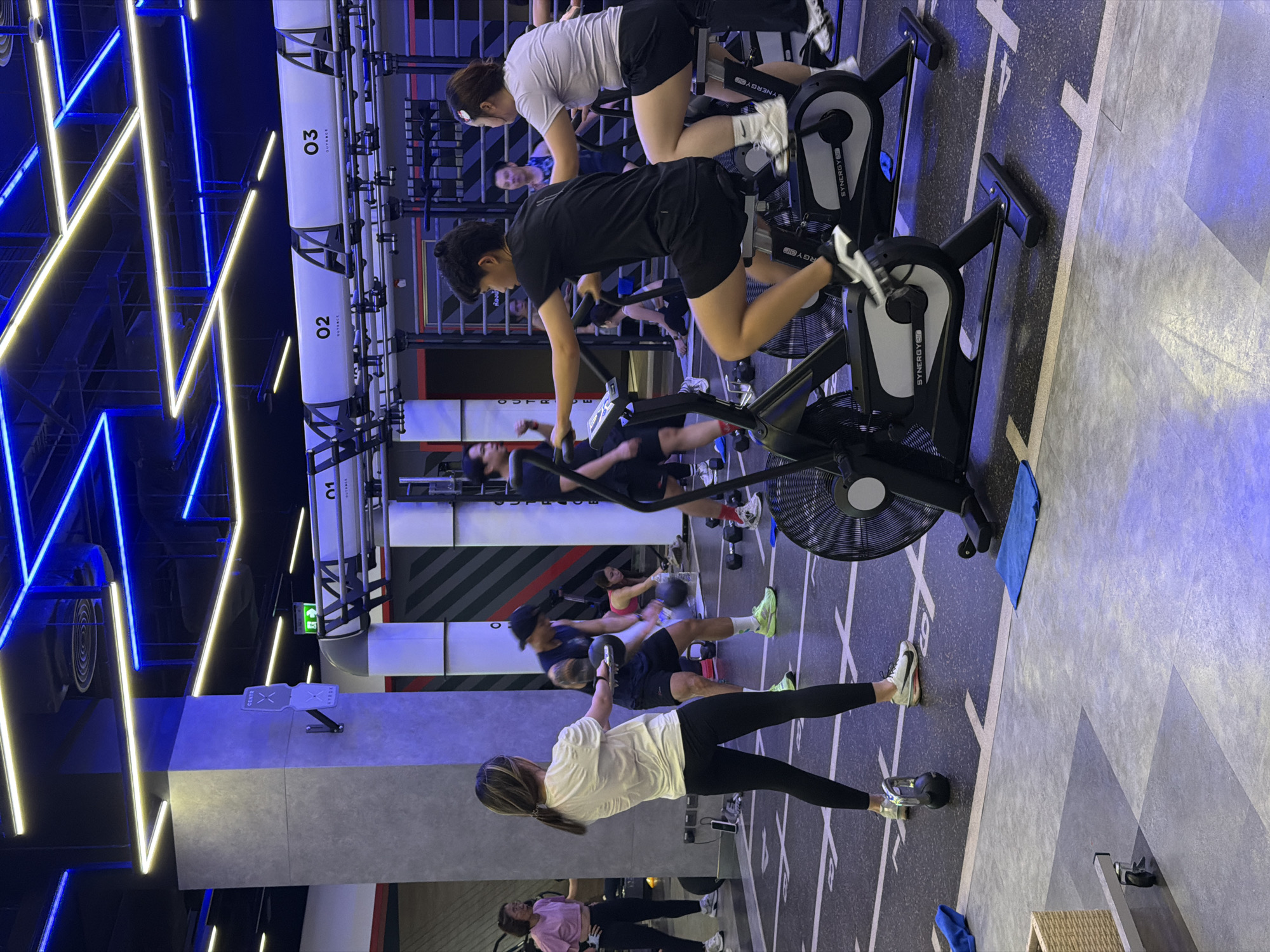 The image shows an indoor gym setting with several people engaged in various exercises. The gym is brightly lit with blue and yellow LED strip lights on the ceiling, creating a modern and energetic atmosphere.
In the foreground, a woman with her back to the camera is performing a kettlebell swing. She is wearing a white t-shirt and black leggings. To her right, a man in a black sleeveless shirt and shorts is also holding a kettlebell, seemingly mid-workout.
Behind them, two individuals are on stationary bikes. The person on the left, wearing a black t-shirt and shorts, is cycling with determination. The person on the right, in a grey t-shirt and black shorts, is also on a bike.
Further in the background, other individuals can be seen exercising. One woman in a pink top and black leggings is standing, and another person in a blue t-shirt is also visible. Dumbbells and other gym equipment are scattered on the floor. The gym floor is marked with white lines, possibly indicating workout zones or lanes.
The overall scene depicts a dynamic and well-equipped fitness facility where people are actively participating in their workouts. The location is identified as Nong Pa Khrang, Thailand.