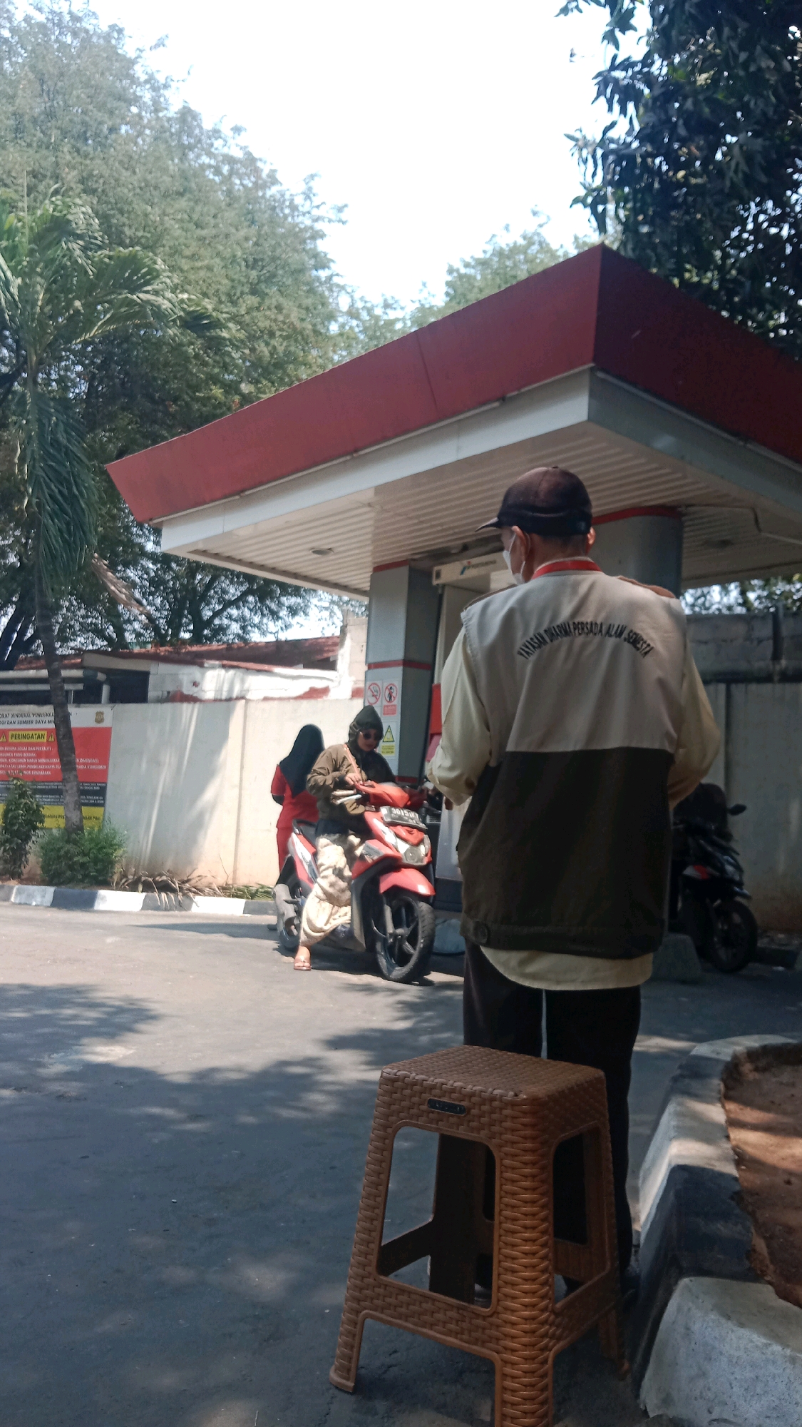 The image depicts an outdoor scene at a gas station in Bekasi, Indonesia. In the foreground, a person wearing a dark baseball cap and a two-toned jacket with writing on the back stands facing away from the camera, with a brown plastic stool to their right. Behind this person, two individuals are on a red motorcycle. One person is operating the motorcycle, while another sits behind them. The gas station canopy, with its red trim, is visible overhead. Trees and foliage surround the area. A wall with signage and a palm tree are also present. The ground is paved asphalt, with sunlight casting shadows. A second motorcycle is parked to the right, partially obscured.