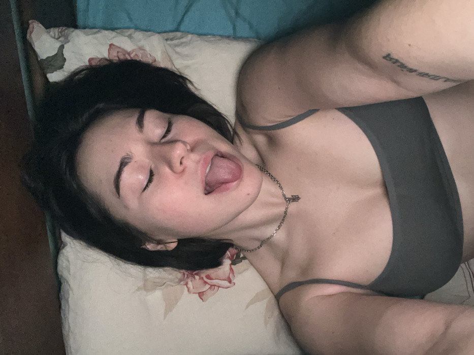 A woman with short dark hair lies on a floral pillow, eyes closed and mouth open, tongue extended. She wears a gray bra top and a silver chain necklace. Her skin appears light-toned. The background consists of a blue sheet and a wooden surface. The angle of the shot is from above, showcasing her face and upper body. Tattoos are visible on her upper arm.